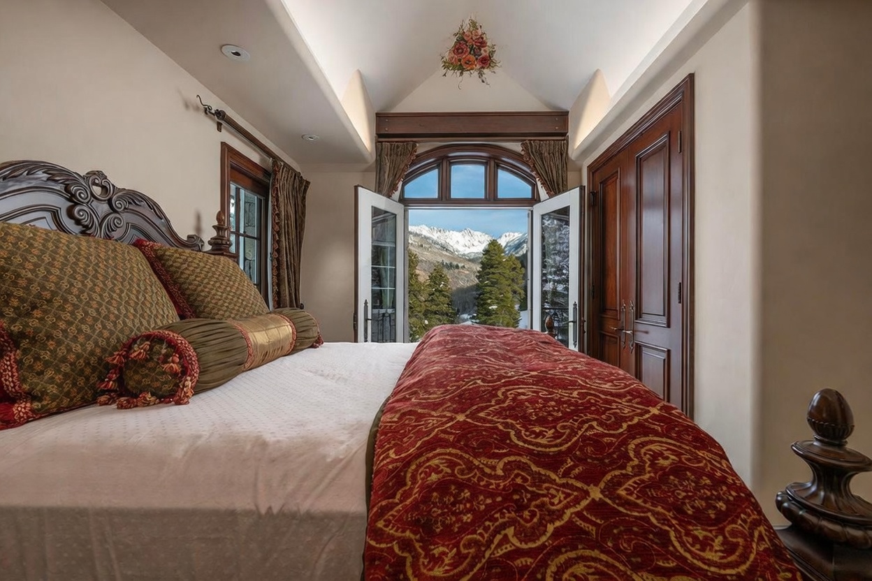 This luxurious primary bedroom features a grand, dark wood bed frame adorned with ornate, patterned bedding and a decorative bolster pillow. The room is highlighted by a pair of open French doors that frame a breathtaking, unobstructed view of snow-capped mountains and evergreen trees. The vaulted ceiling includes a decorative floral accent, and the warm, rich wood tones of the doors and furniture create a sophisticated and cozy mountain retreat atmosphere.