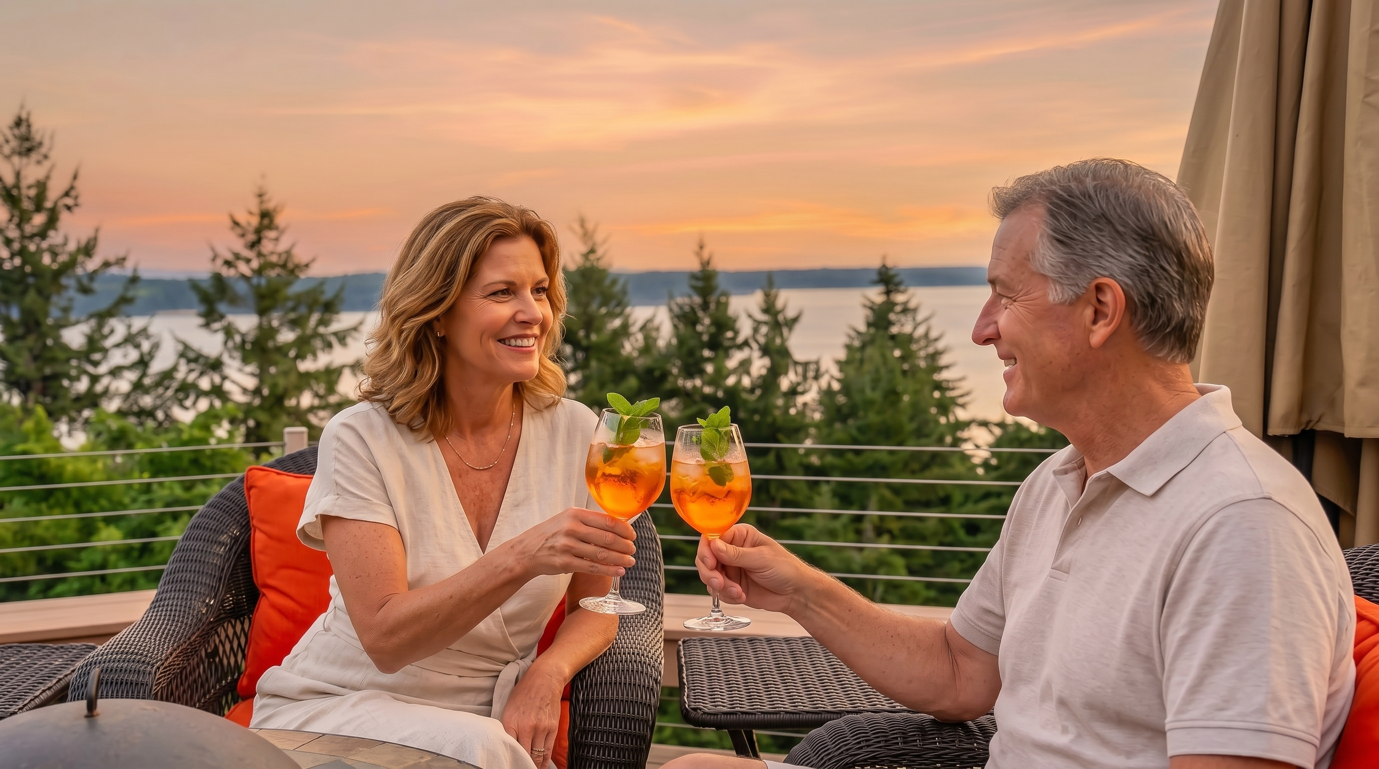This expansive outdoor deck offers a stunning panoramic view of the water and surrounding evergreen forest. The space is furnished with a circular fire pit surrounded by comfortable wicker seating with vibrant orange cushions, creating an inviting area for relaxation. A large, closed patio umbrella stands to the side, and the deck is enclosed by a modern cable railing system that preserves the scenic vista.