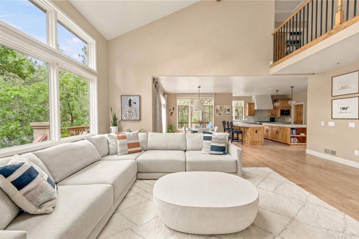 This spacious, open-concept living area features a large, light-colored sectional sofa arranged on a textured area rug, centered around a low, round coffee table. The room boasts high, vaulted ceilings and expansive windows that provide abundant natural light and views of the surrounding trees. Beyond the living space, the layout flows seamlessly into a kitchen area with a large island and bar seating, creating an airy and inviting atmosphere.