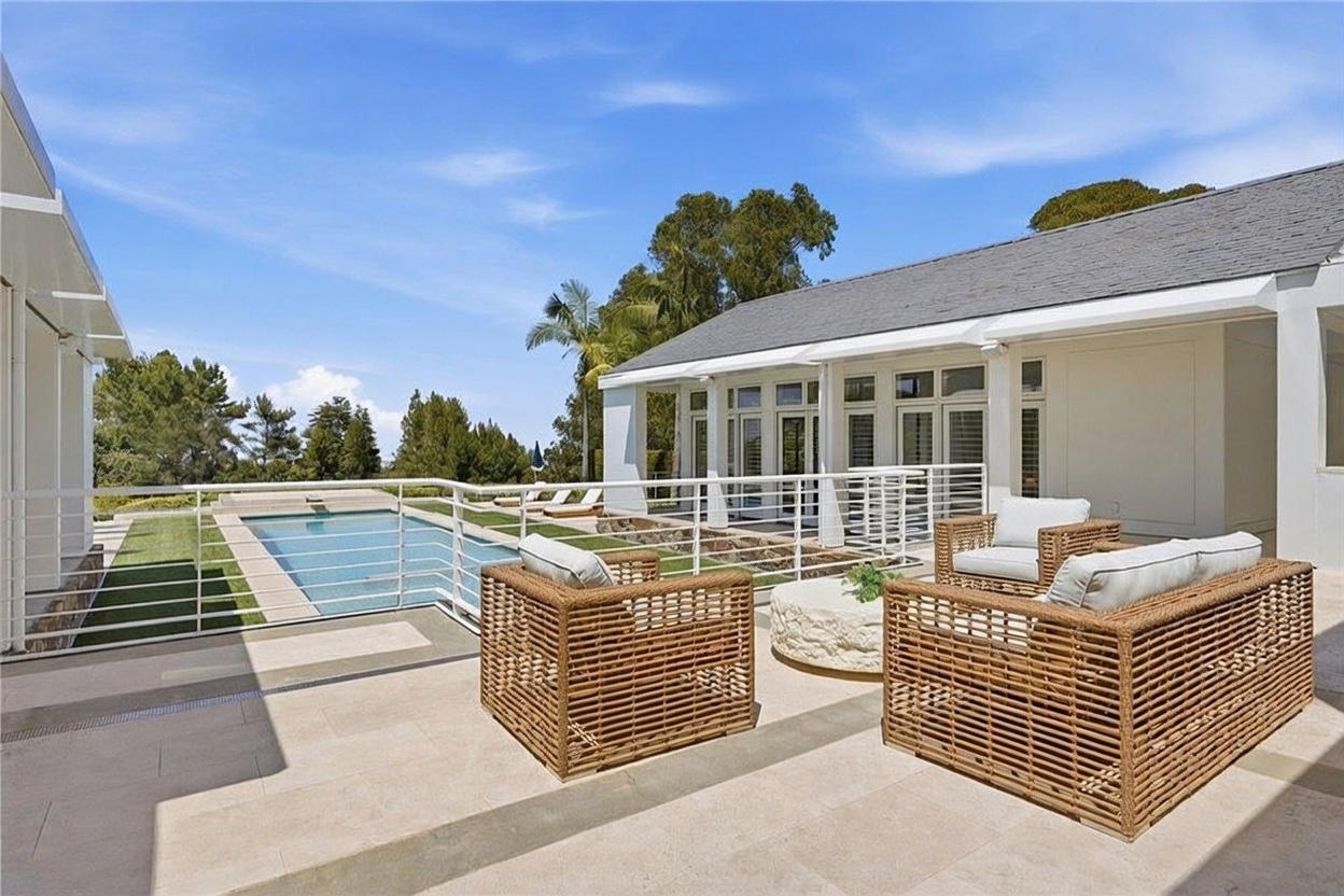 This bright outdoor patio space features woven wicker-style armchairs arranged around a low, circular stone table, creating a comfortable seating area. The patio is paved with light-toned stone tiles and overlooks a rectangular swimming pool surrounded by a white metal railing. The scene is framed by a white-walled structure with large glass doors and windows, set against a clear blue sky and lush green trees in the background.
