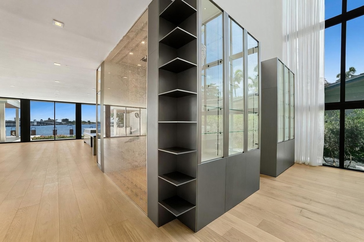 This modern, open-concept living space features light wood flooring and a central, custom-built shelving unit with glass-fronted display cabinets. The room is framed by expansive, floor-to-ceiling windows that provide a clear view of the waterfront outside. The minimalist design emphasizes clean lines and a bright, airy atmosphere, creating a sophisticated and seamless transition between the indoor living area and the exterior landscape.
