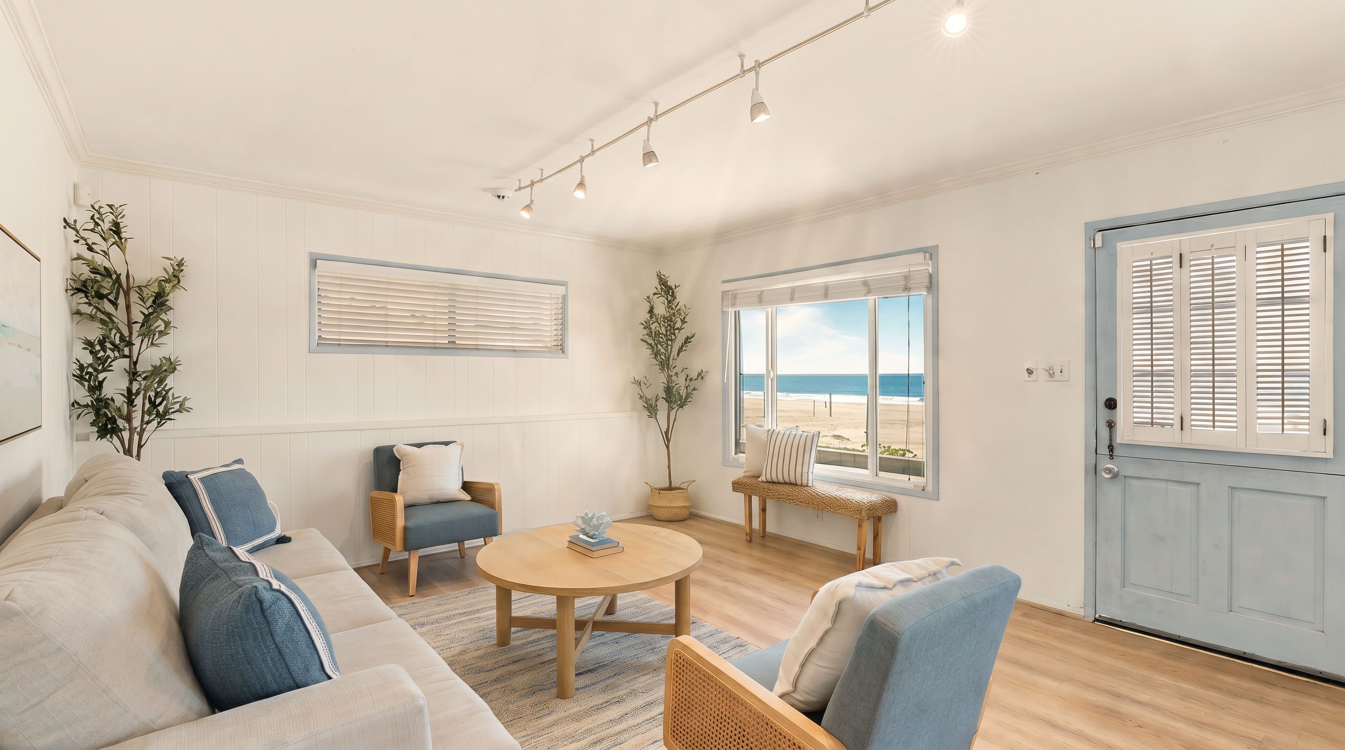 This bright and airy living room features a coastal-inspired aesthetic with light wood-tone flooring and white paneled walls. The space is furnished with a neutral sofa, two blue accent chairs, and a round wooden coffee table, all arranged on a textured area rug. A large window provides a direct, scenic view of the beach and ocean, while track lighting mounted on the ceiling illuminates the room.