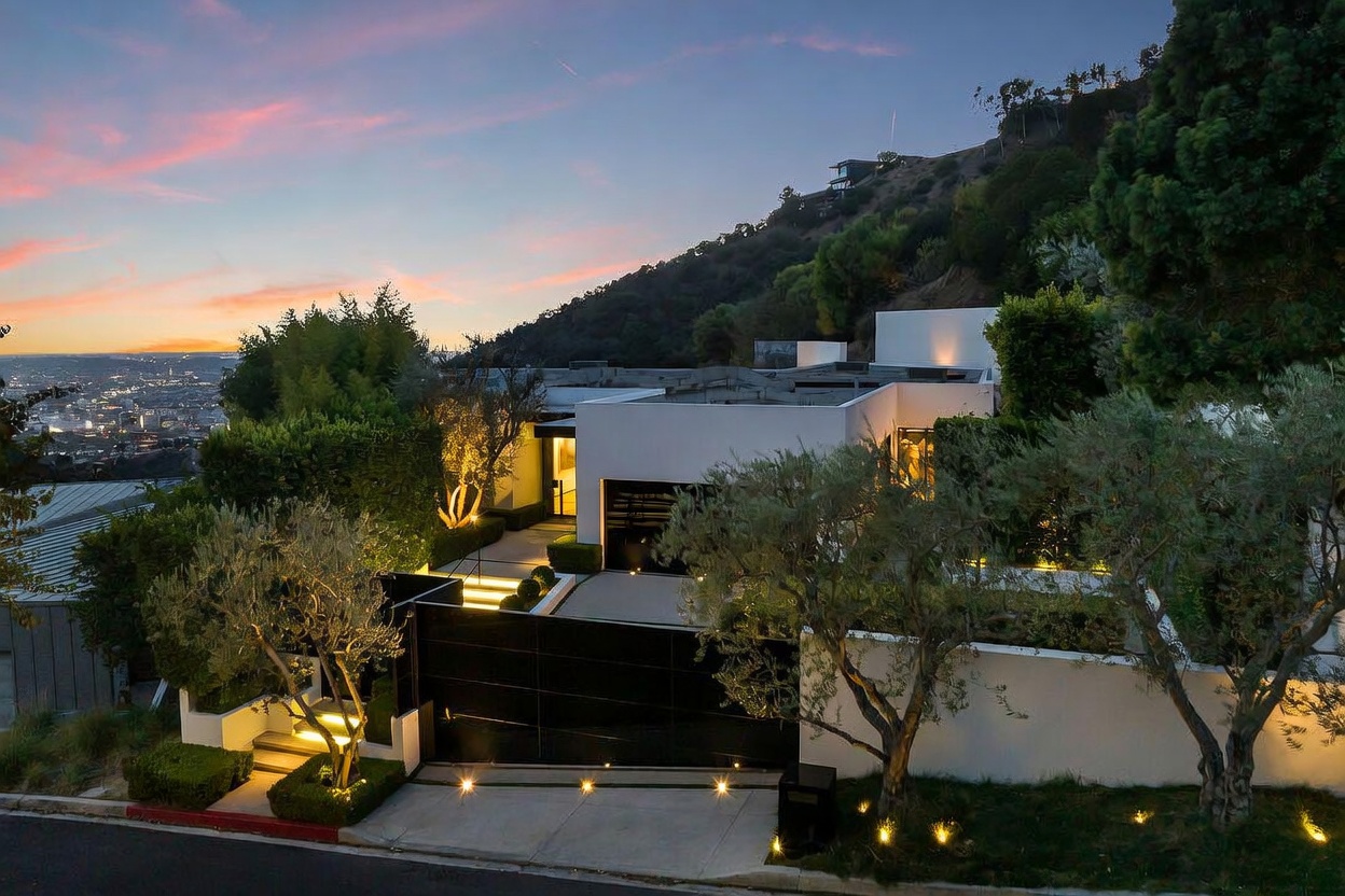 This modern, minimalist residence is captured from an elevated street-level perspective at dusk, showcasing its sleek white geometric architecture against a dramatic sunset sky. The property features a prominent black gated entrance, manicured landscaping with olive trees, and warm integrated lighting that highlights the driveway and pathways. The home is nestled into a hillside, offering a commanding view of the sprawling city lights in the distance.