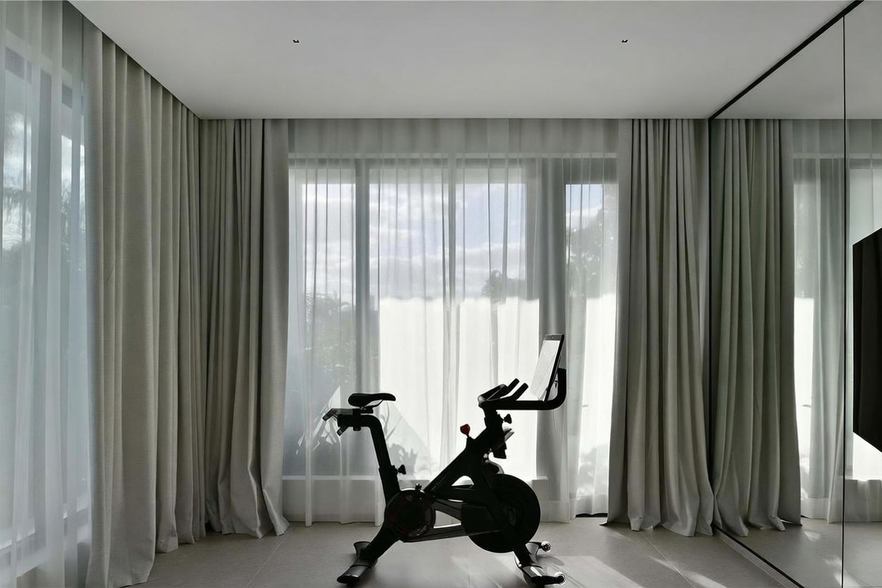 This minimalist home gym features a stationary exercise bike positioned centrally in front of large, floor-to-ceiling windows draped in soft, neutral-toned curtains. The room is finished with light-colored tile flooring and a large mirror on the right wall, which enhances the sense of space and light. The overall aesthetic is clean, modern, and serene, providing an inviting atmosphere for a workout.