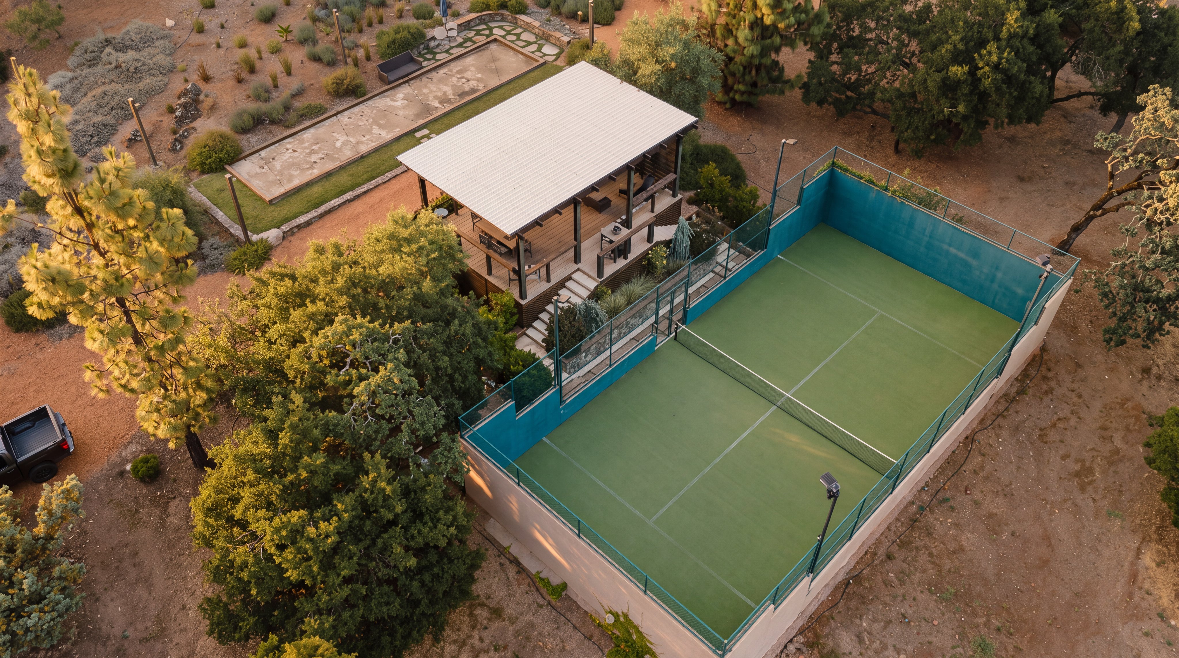 This high-angle aerial view showcases a well-appointed outdoor recreational area featuring a fenced tennis court with a green surface. Adjacent to the court is a covered pavilion with a metal roof, providing a shaded space for relaxation, and a long bocce ball court set into the landscaped grounds. The property is surrounded by mature trees and natural terrain, creating a private and serene environment for outdoor activities.