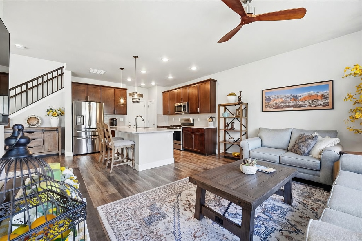 This open-concept living space features a kitchen with dark wood cabinetry, stainless steel appliances, and a central island with bar seating. The adjacent living area is furnished with a light-colored sofa and a rustic coffee table, all set upon a patterned area rug. The room is illuminated by recessed lighting and a ceiling fan, creating a warm and inviting atmosphere with a clear view of the staircase and upper level.