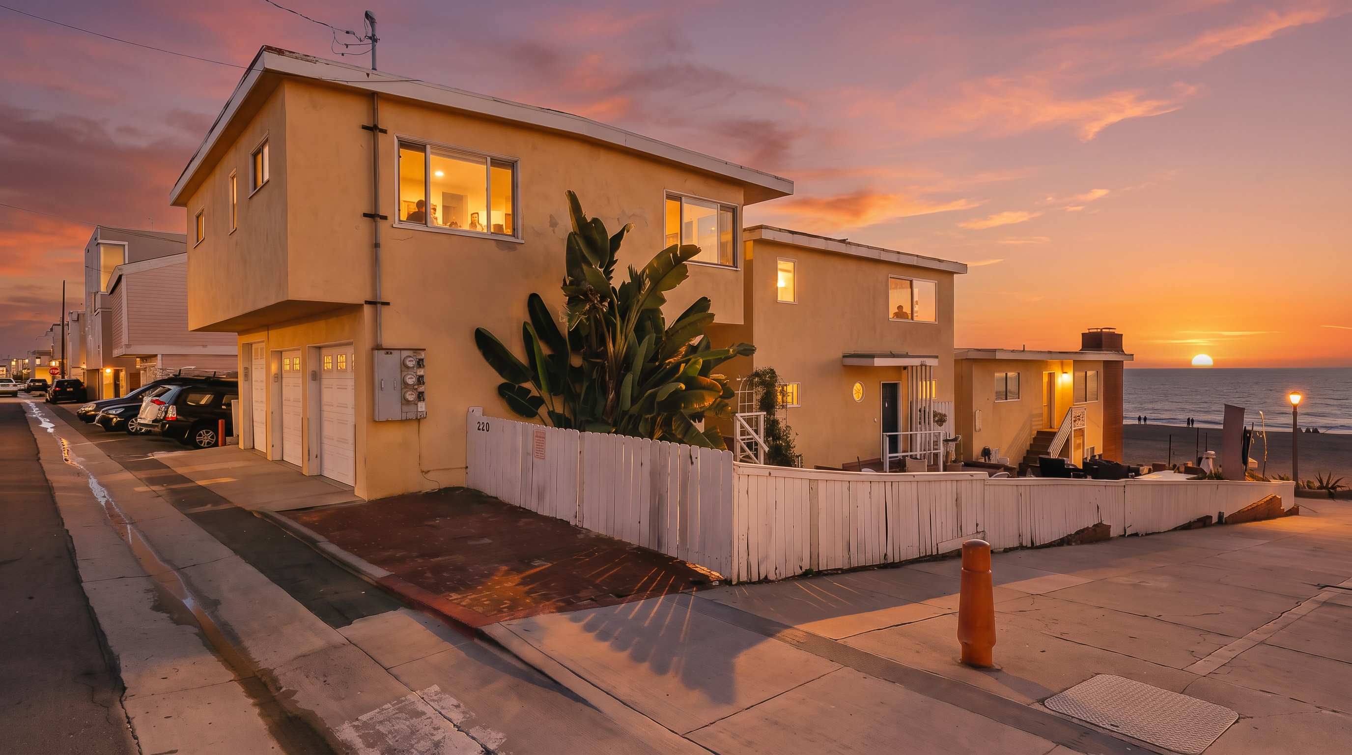 This exterior view captures a multi-level, light-yellow stucco property situated directly adjacent to the beach. The structure features a series of garage doors on the ground level, a white picket fence enclosing a small yard area, and large windows that offer views of the ocean. The perspective is taken from the street level, highlighting the property's prime coastal location and its proximity to the sandy shoreline.