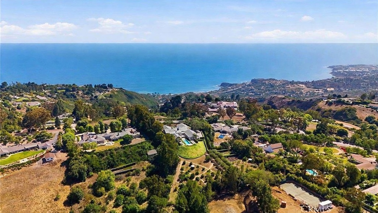 This high-altitude aerial view captures a sprawling estate nestled on a hillside overlooking the vast Pacific Ocean. The property features a main residence with a swimming pool, surrounded by extensive landscaped grounds, mature trees, and a private tennis court. The perspective emphasizes the expansive coastal location and the secluded, elevated nature of the home.