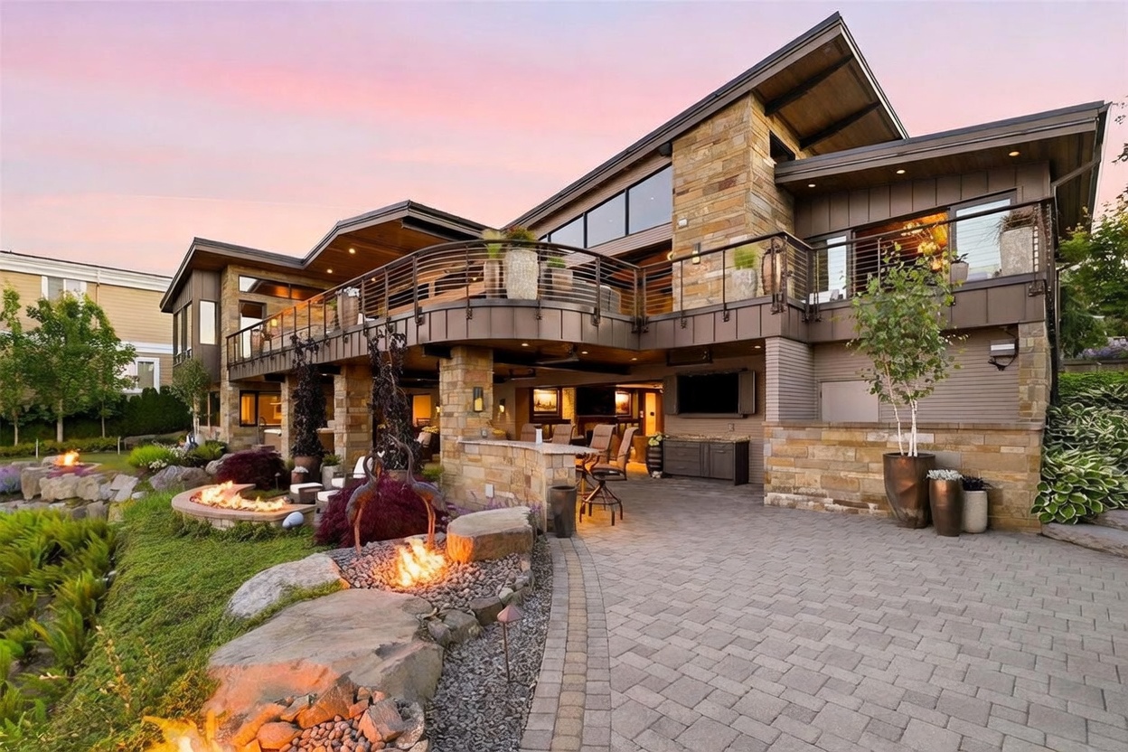 This stunning contemporary residence features a multi-level rear exterior with a spacious stone-paved patio and a curved upper-level balcony. The home showcases a sophisticated blend of stone masonry and dark metal siding, complemented by large windows and an integrated outdoor kitchen area. The landscaped yard includes a fire pit set within natural rock formations, creating a luxurious and inviting atmosphere for evening entertaining.