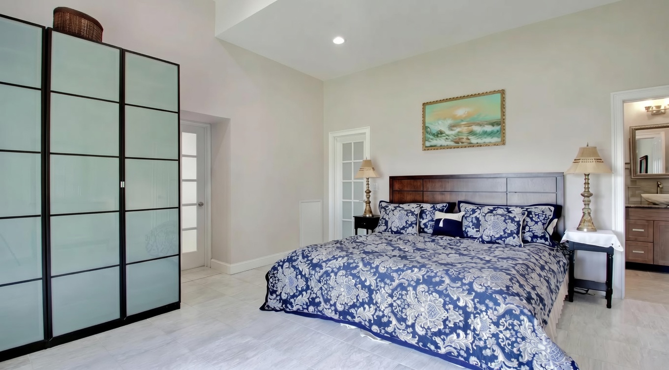 This spacious primary bedroom features neutral-toned walls, high vaulted ceilings, and light-colored tile flooring, creating an airy and open atmosphere. It is furnished with a large bed topped with a bold blue and white patterned comforter, flanked by classic bedside lamps and a contemporary floor-to-ceiling wardrobe unit. The presence of multiple doorways, including a transition to an en-suite bathroom, adds convenience and highlights the functional layout of the room.