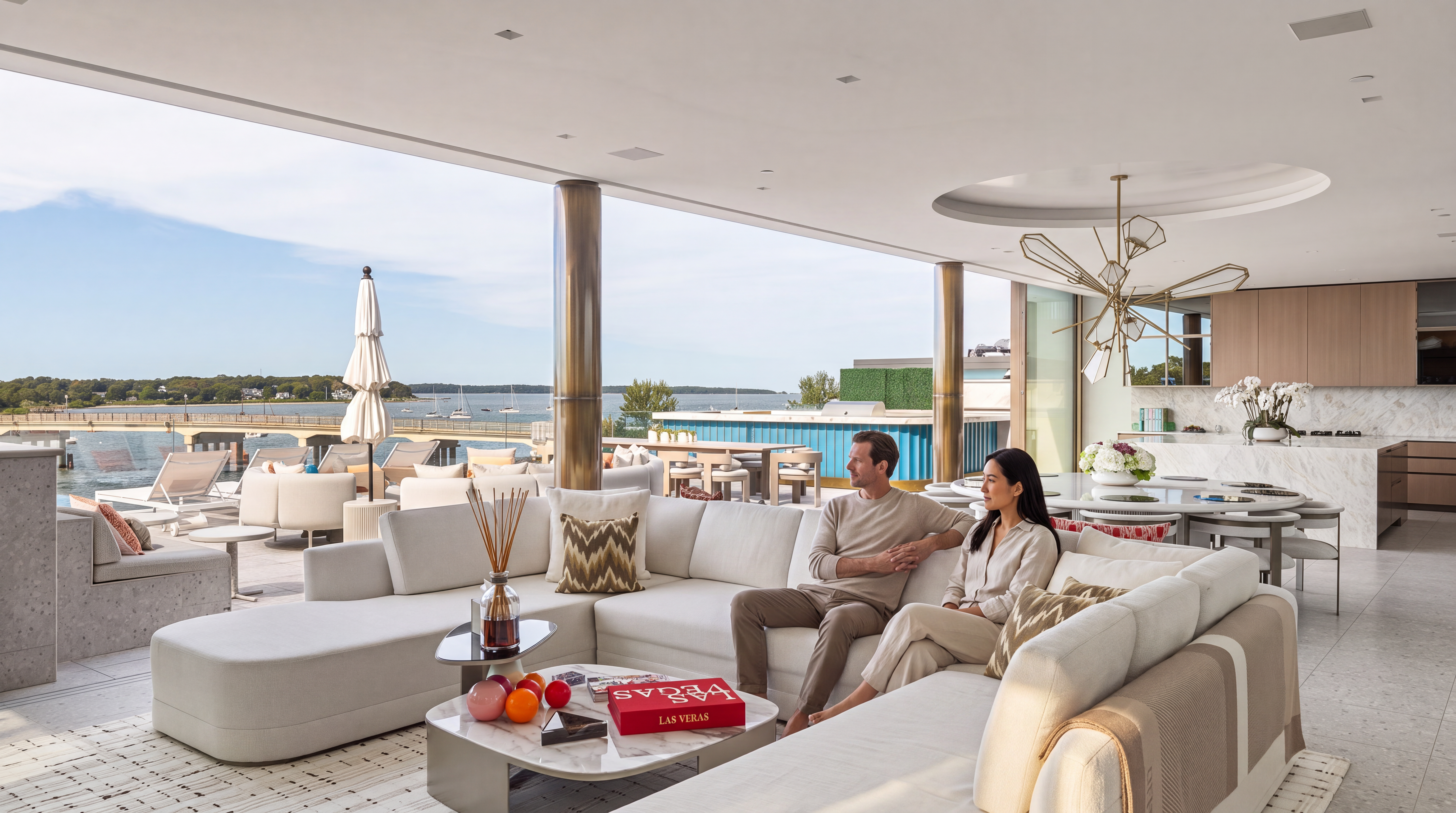 This luxurious open-concept living area seamlessly transitions to a spacious outdoor deck overlooking the water through expansive glass walls. The room features a plush ivory sectional, a modern marble-topped coffee table, and an adjacent high-end kitchen, creating an inviting space for relaxation and entertaining. The sophisticated design includes a unique sculptural chandelier and elegant neutral tones, emphasizing a contemporary indoor-outdoor lifestyle.