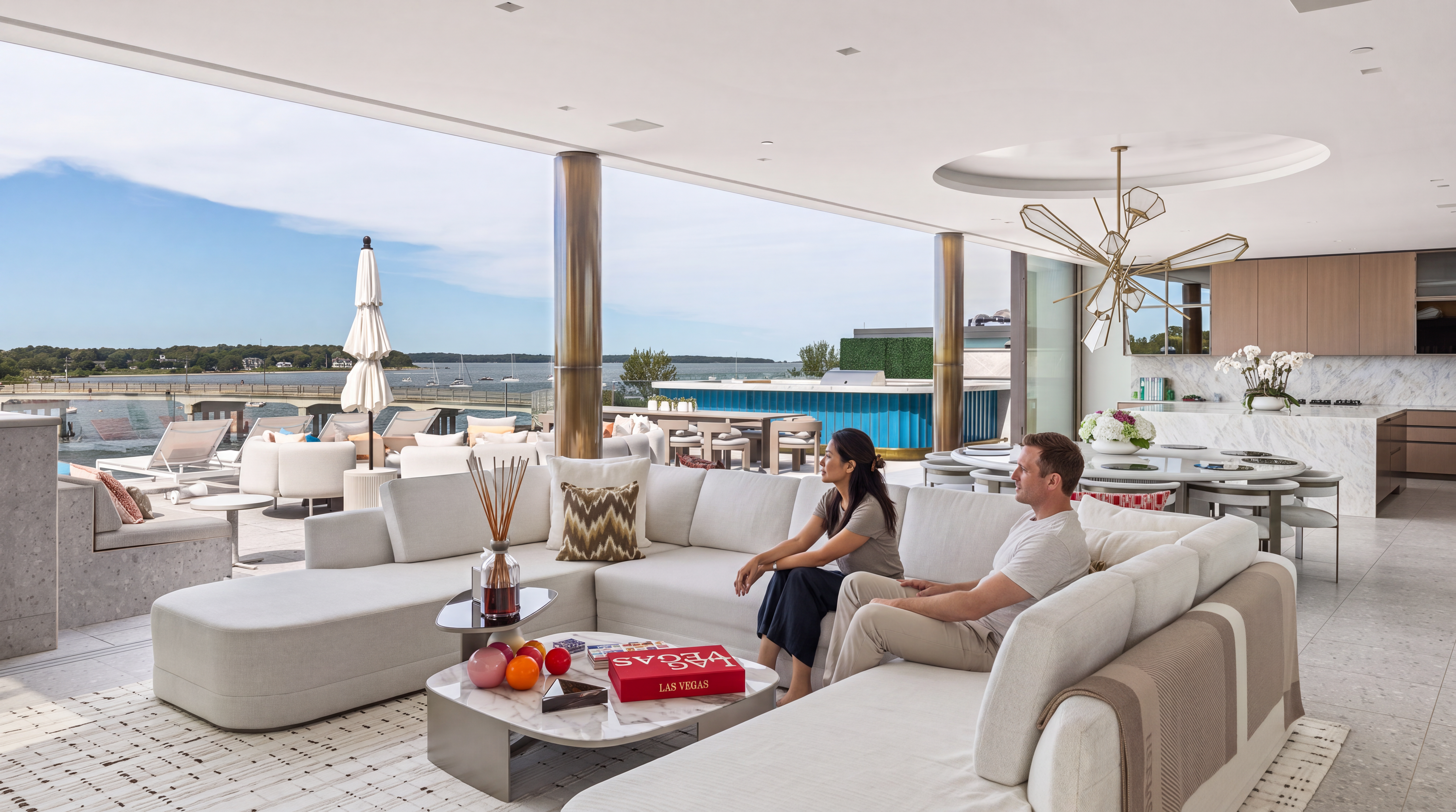 This luxurious, open-concept living space seamlessly integrates with a expansive outdoor terrace, featuring floor-to-ceiling glass and panoramic water views. The interior boasts a contemporary aesthetic with a large, comfortable sectional sofa, high-end marble coffee table, and a bespoke chandelier that serves as a striking centerpiece. The design emphasizes indoor-outdoor living, making it an ideal setting for entertaining in a serene, waterfront environment.