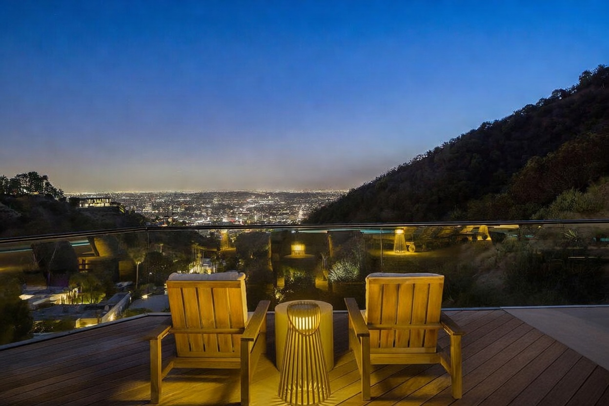 This elevated wooden deck offers a panoramic, cinematic view of a sprawling city skyline at twilight. Two light-toned wooden chairs are positioned around a cylindrical, slatted lantern-style light fixture, creating an intimate seating area behind a sleek glass railing. The scene is illuminated by warm, ambient lighting that contrasts beautifully with the deep blue evening sky and the distant urban glow.