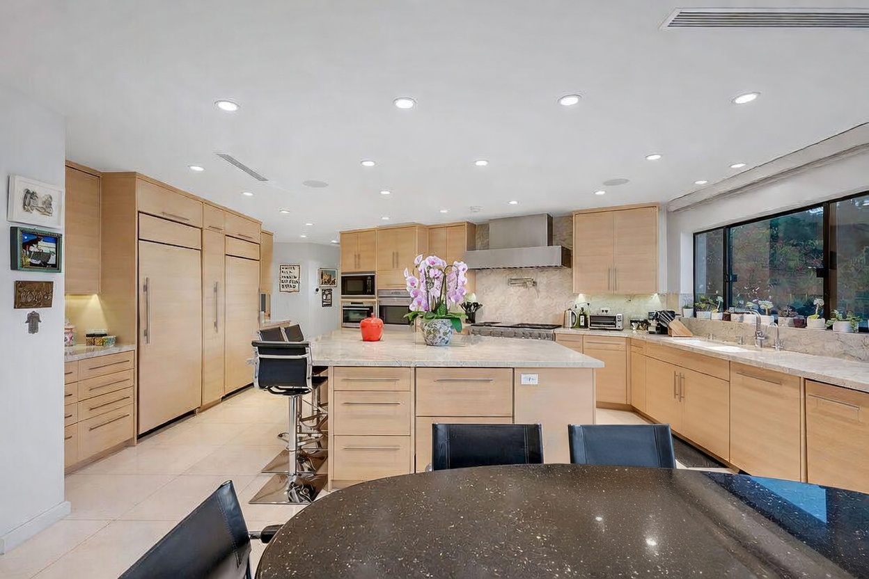 This spacious, modern kitchen features light wood cabinetry, a large central island with stone countertops, and integrated stainless steel appliances. A circular dining table with a dark surface sits in the foreground, accented by a vibrant orchid arrangement, while recessed lighting illuminates the entire open-concept space. The perspective captures the functional layout, highlighting the seamless transition between the food preparation area and the casual dining zone.
