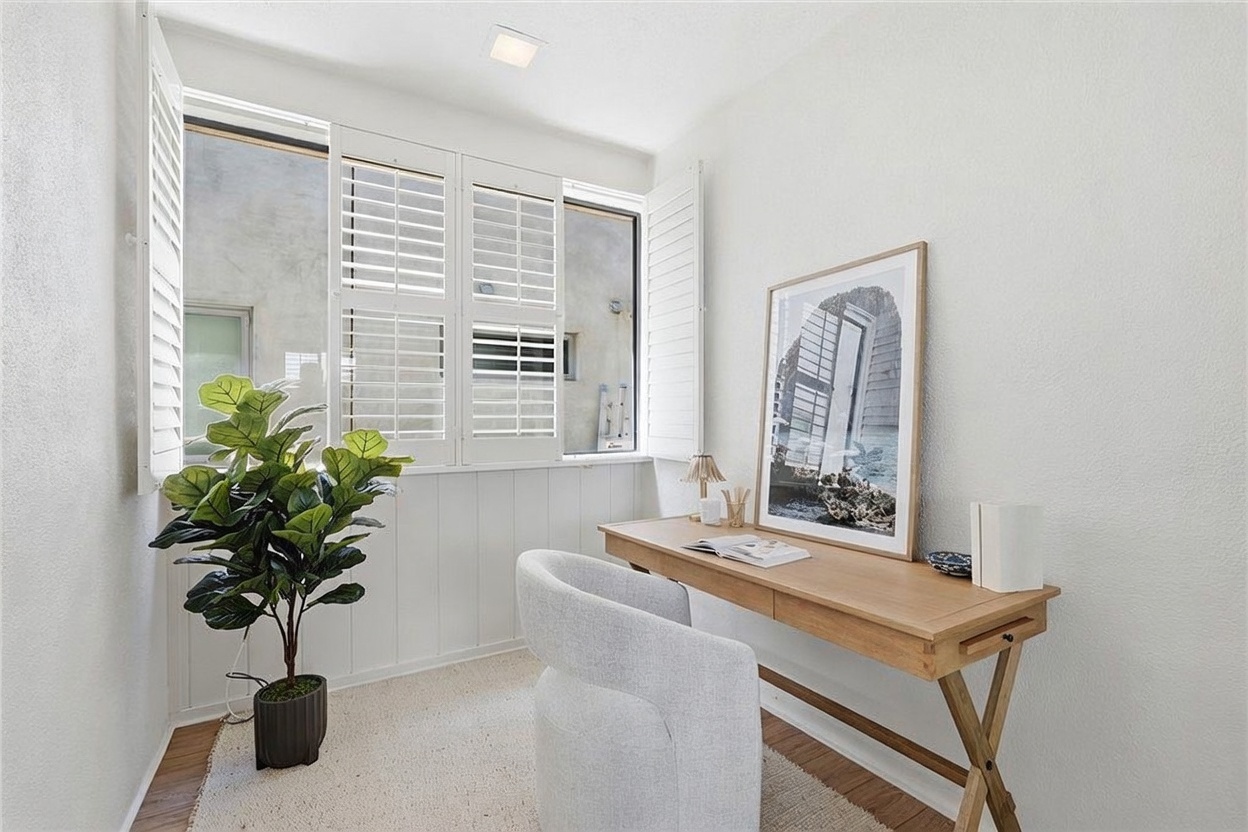 This bright and airy home office features a minimalist aesthetic with white walls and light wood flooring. A wooden desk is positioned against the wall, complemented by a modern, curved accent chair and a large framed piece of art. The space is illuminated by natural light streaming through a set of windows adorned with white plantation shutters, while a potted plant adds a touch of greenery to the corner.