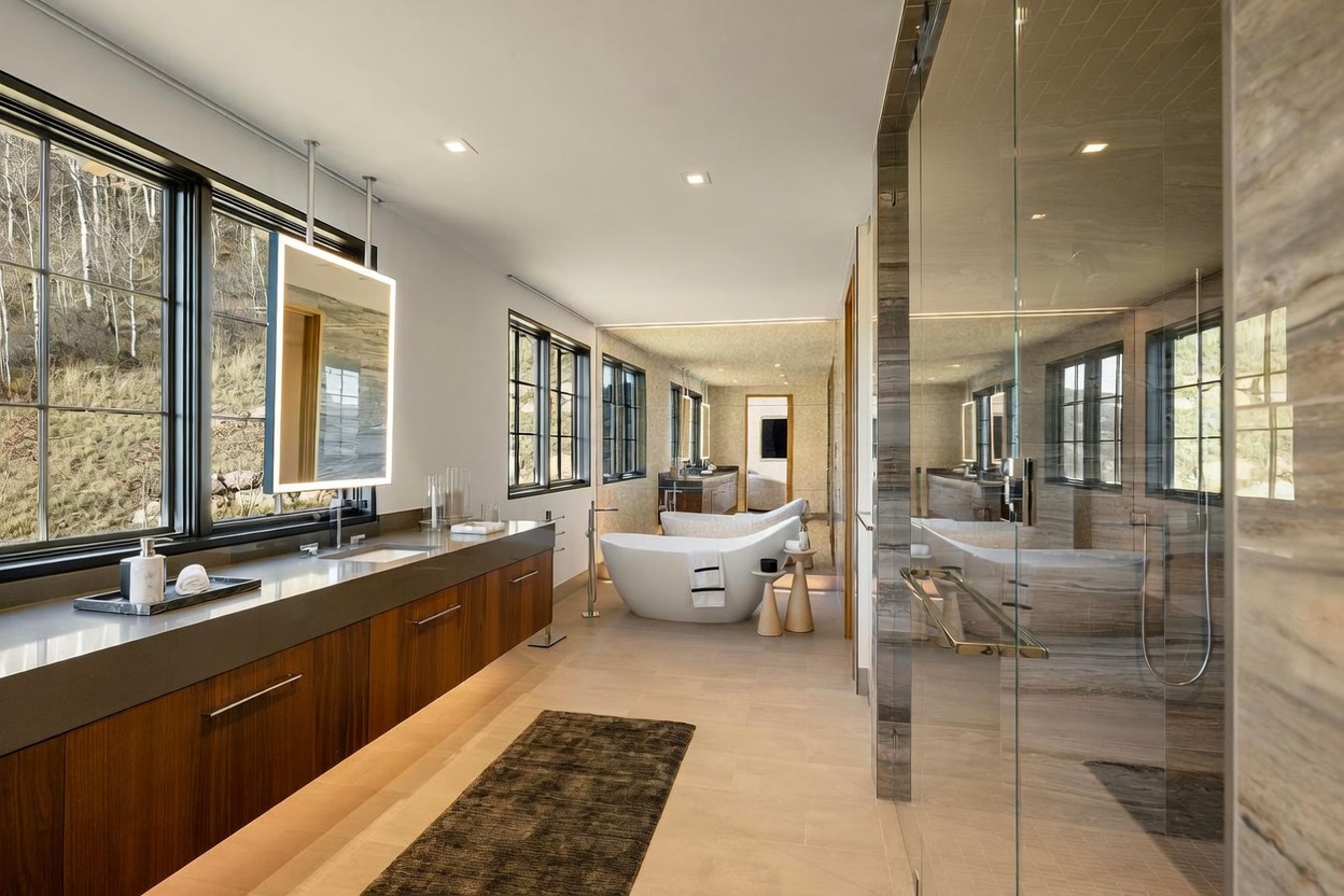 This luxurious primary bathroom features a long, floating vanity with a dark wood finish and a sleek stone countertop, paired with a modern, illuminated mirror. A freestanding soaking tub serves as a focal point in the center of the room, complemented by a glass-enclosed walk-in shower on the right. Large windows along the left wall provide ample natural light and views of the outdoors, creating an open and serene atmosphere.