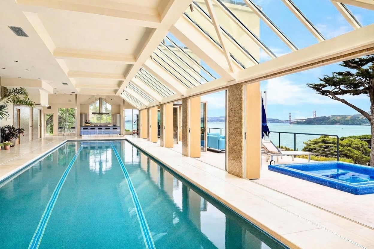 This luxurious indoor-outdoor lap pool area features a long, rectangular pool with a dedicated lane marker, adjacent to a raised, tiled spa. The space is enclosed by a dramatic glass-paneled roof and retractable doors that open to a stunning panoramic view of the bay and the Golden Gate Bridge. The design emphasizes a seamless transition between the climate-controlled interior and the natural beauty of the coastal landscape.