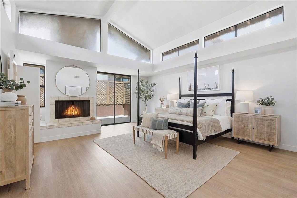 This spacious primary bedroom features a dramatic vaulted ceiling with clerestory windows that flood the room with natural light. A black four-poster bed serves as the focal point, complemented by a cozy fireplace with a light-toned tile surround and a large circular mirror above. The room is finished with light wood-look flooring and a neutral color palette, creating an airy and sophisticated atmosphere.