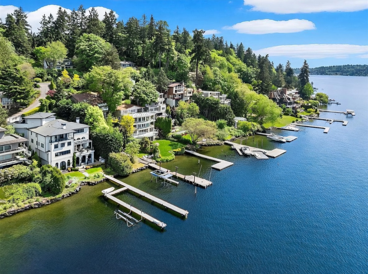 This high-angle aerial view captures a row of luxury waterfront homes nestled along a lush, tree-lined hillside overlooking a calm lake. Each property features private docks extending into the water, with several boats moored at the piers. The scene highlights the seamless integration of upscale residential architecture with the natural landscape, emphasizing the serene and exclusive nature of the waterfront lifestyle.