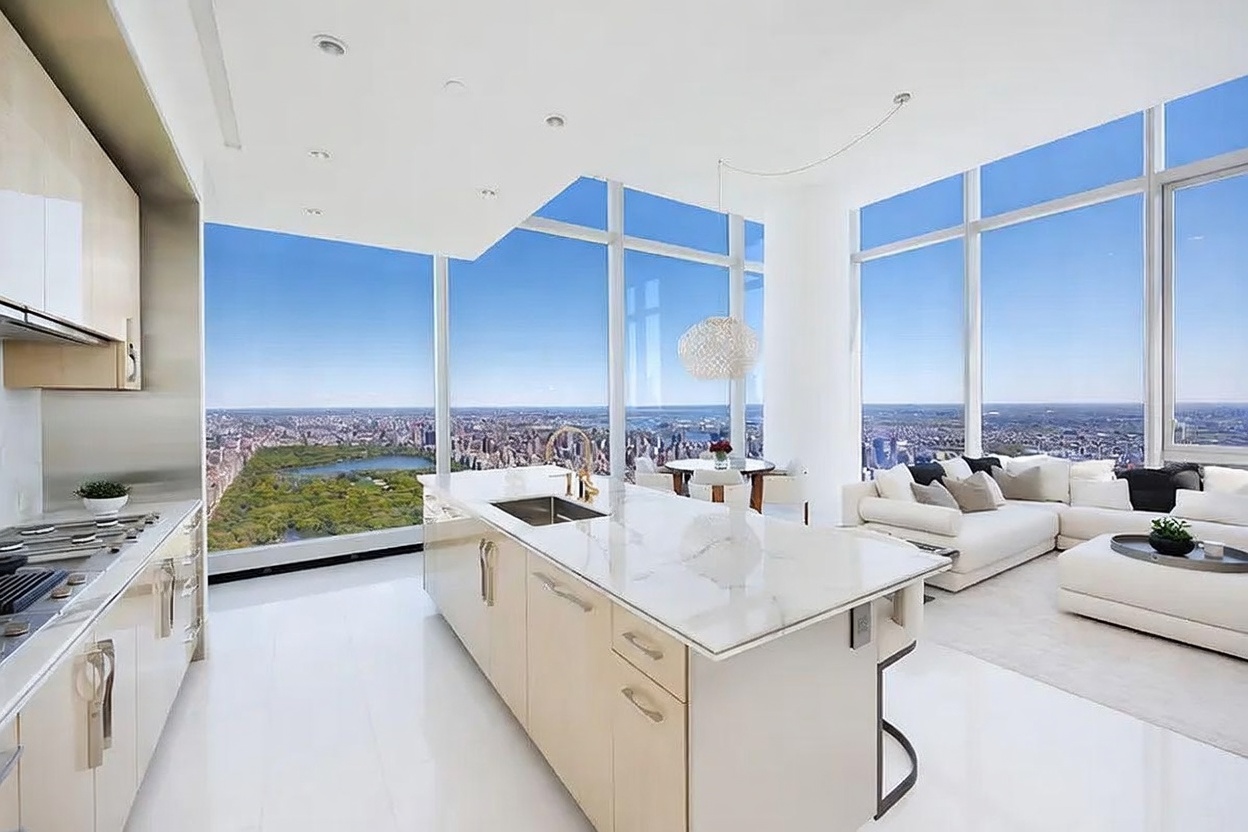 This high-rise kitchen features a sleek, minimalist design with light wood cabinetry and a large central island topped with a light-toned stone surface. The space is defined by expansive floor-to-ceiling windows that provide a dramatic, panoramic view of Central Park and the surrounding city skyline. Adjacent to the kitchen, a bright living area with white seating and a dining nook creates an open-concept layout bathed in natural light.
