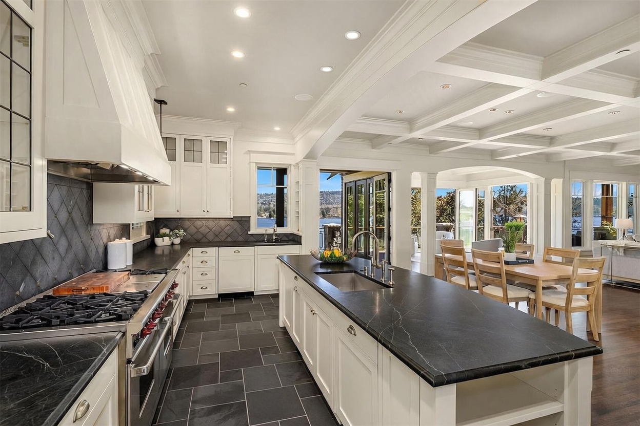 This elegant, high-end kitchen features a spacious island with a dark stone countertop, white cabinetry, and professional-grade stainless steel appliances. The room is accented by a coffered ceiling, dark slate flooring, and a sophisticated backsplash, all bathed in natural light from large windows that offer a scenic view of the water. The open-concept design seamlessly connects the kitchen to the dining area, creating a bright and inviting atmosphere perfect for entertaining.