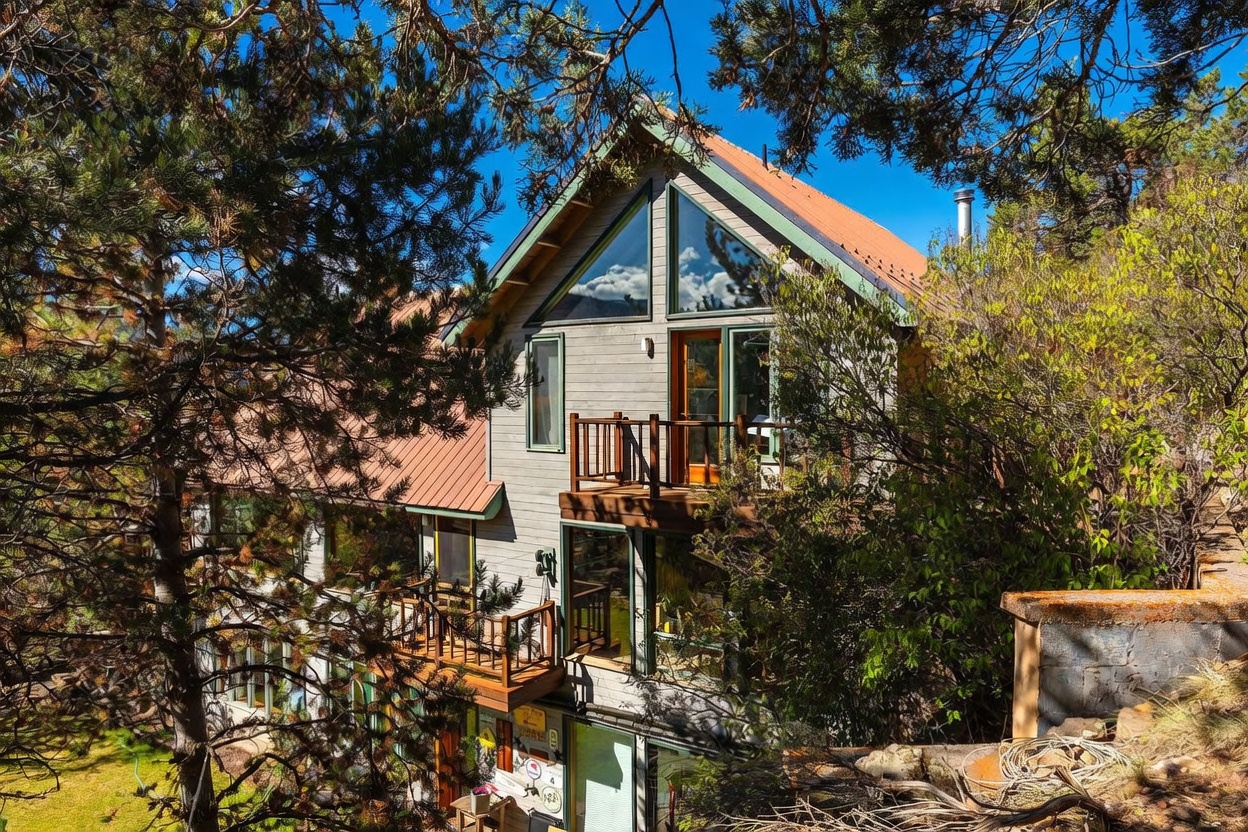 This multi-level mountain home features a rustic aesthetic with light-toned wood siding, a metal roof, and large triangular windows that capture the surrounding forest. The structure includes multiple wooden decks and balconies that provide outdoor living space nestled among mature pine trees. The perspective is slightly elevated, showcasing the home's integration into the sloped, wooded landscape.