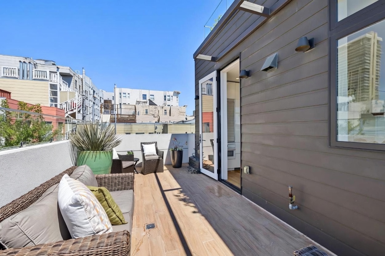 This modern rooftop deck features wood-look tile flooring and a comfortable outdoor seating area with wicker furniture. The space is enclosed by a glass railing, offering an open view of the surrounding urban neighborhood, and connects directly to the interior through a glass door. The clean lines and contemporary design create a bright, inviting outdoor living space perfect for relaxation.