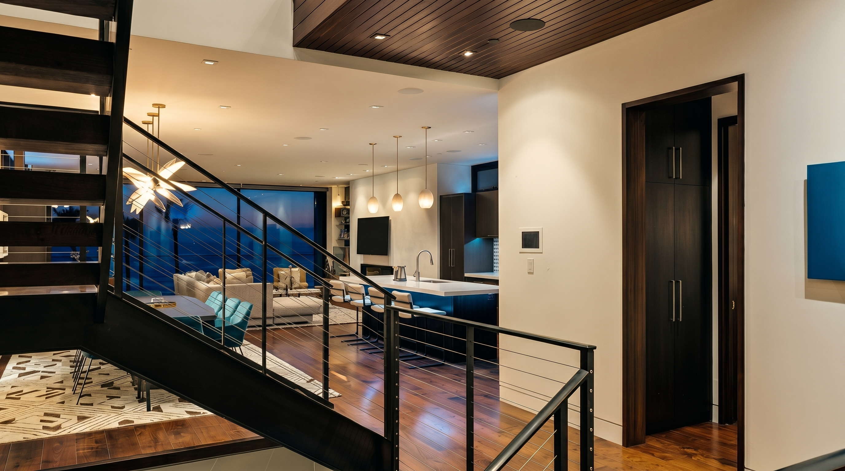 This perspective from a staircase landing overlooks an open-concept living and kitchen area, showcasing a modern, multi-level design. The space features dark wood flooring, a slatted wood ceiling detail, and a sleek kitchen island with bar seating. Large windows at the rear provide a view of the evening sky, while a dark-toned door leads to a storage area on the right.