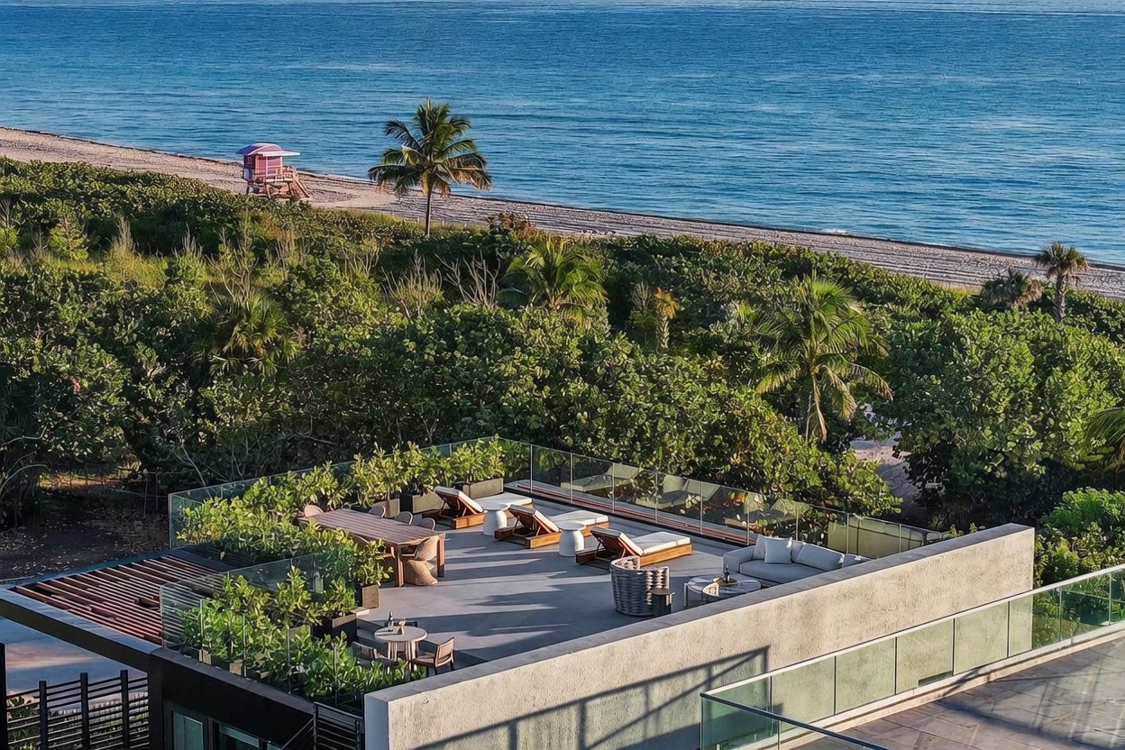 This elevated rooftop terrace offers a luxurious outdoor living space with panoramic views of the ocean and beach. The deck is furnished with a dining set, multiple lounge chairs, and a comfortable sofa, all set against a backdrop of lush tropical greenery. The modern glass railing provides an unobstructed view of the coastline, creating a serene and sophisticated atmosphere.