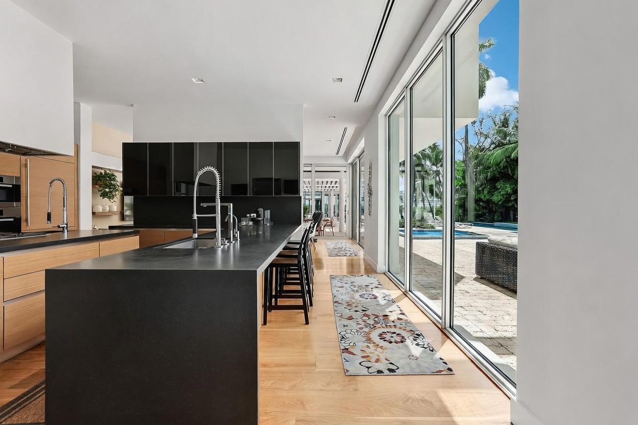 This modern kitchen features a long, dark stone island with bar seating and a sleek, minimalist aesthetic. The space is defined by high-contrast black cabinetry and light wood flooring, with floor-to-ceiling glass sliding doors that provide a seamless transition to the outdoor pool area. The perspective captures the length of the kitchen, emphasizing the open-concept layout and the bright, natural light flooding in from the patio.