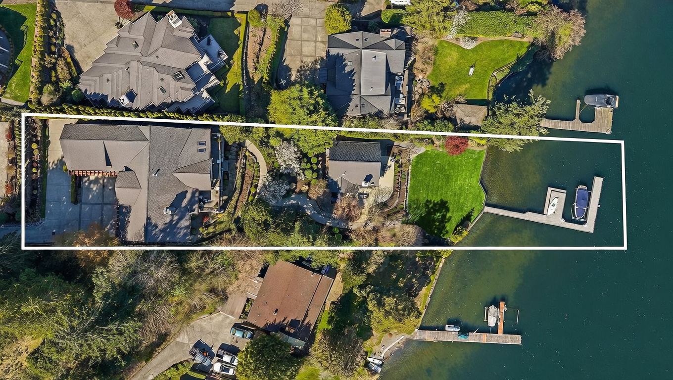 This high-angle aerial view captures a luxurious waterfront property, highlighted by a white boundary outline. The estate features a large main residence, a secondary structure, a lush green lawn, and a private dock extending into the water with two boats moored. The perspective provides a comprehensive look at the property's layout, landscaping, and its prime location along the shoreline.
