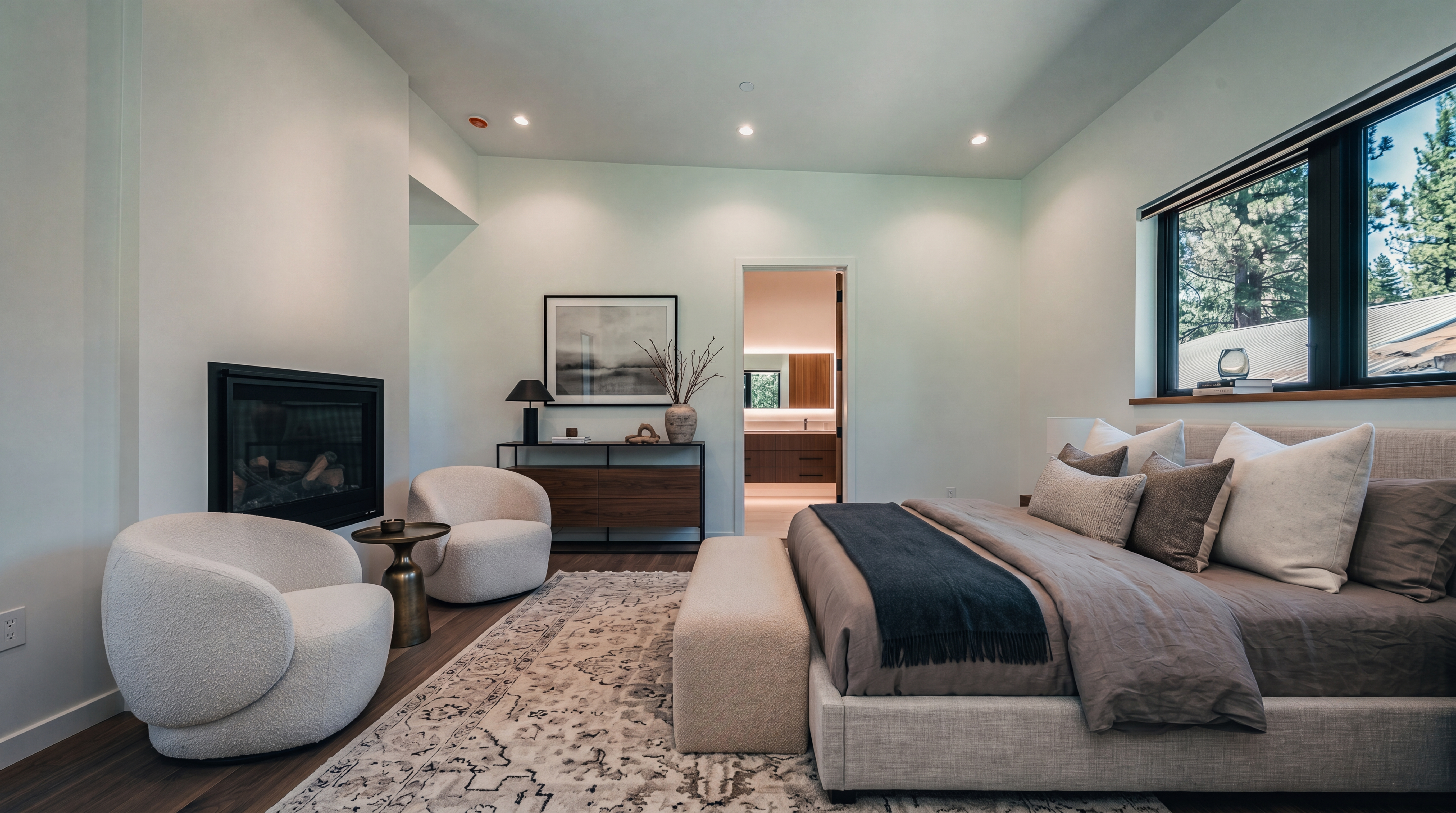 This bright and minimalist bedroom features warm-toned wood flooring and crisp white walls, creating an airy and inviting atmosphere. A black-framed fireplace is built into the wall on the left, while a large window on the right provides natural light and a view of the outdoors. A doorway in the center leads into an adjacent bathroom, showcasing a clean, modern design with a neutral color palette.