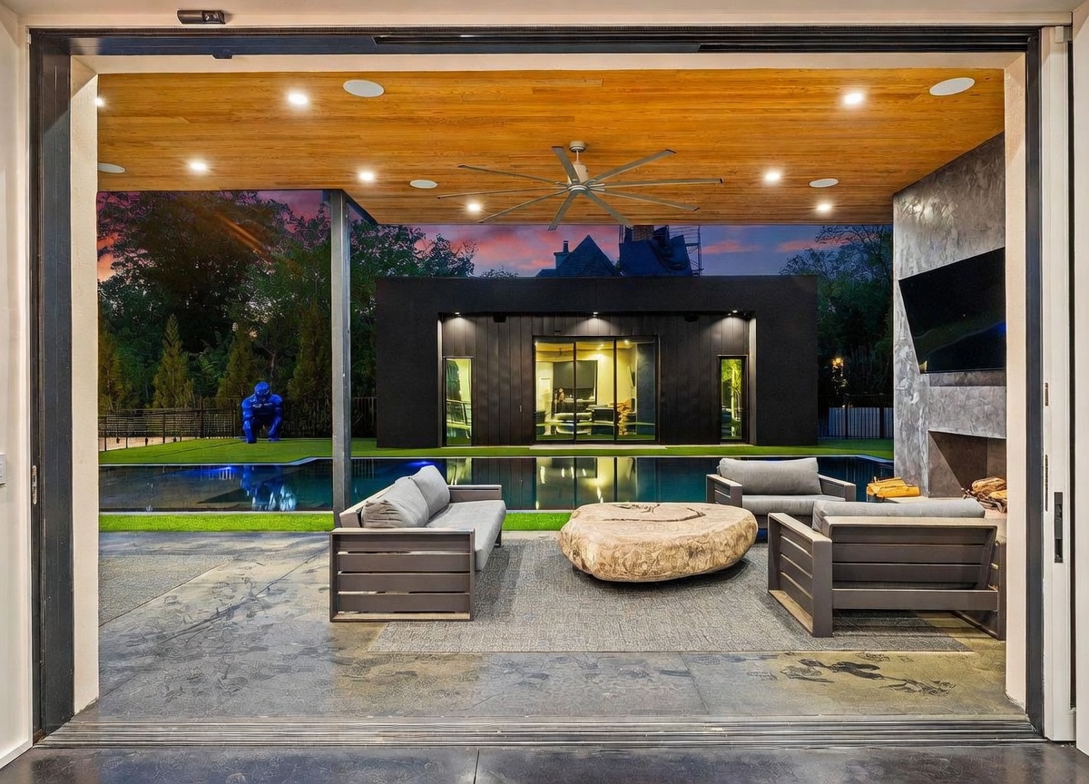 This covered outdoor living space offers a seamless transition from the interior through a wide opening, featuring a wood-paneled ceiling with recessed lighting and a large ceiling fan. The area is furnished with modern lounge seating arranged around a unique, organic-shaped stone coffee table, facing a stone-clad chimney breast with an integrated television. Beyond the patio, a sleek swimming pool reflects the twilight sky, set against a backdrop of a modern black structure and a blue gorilla sculpture on the lawn.