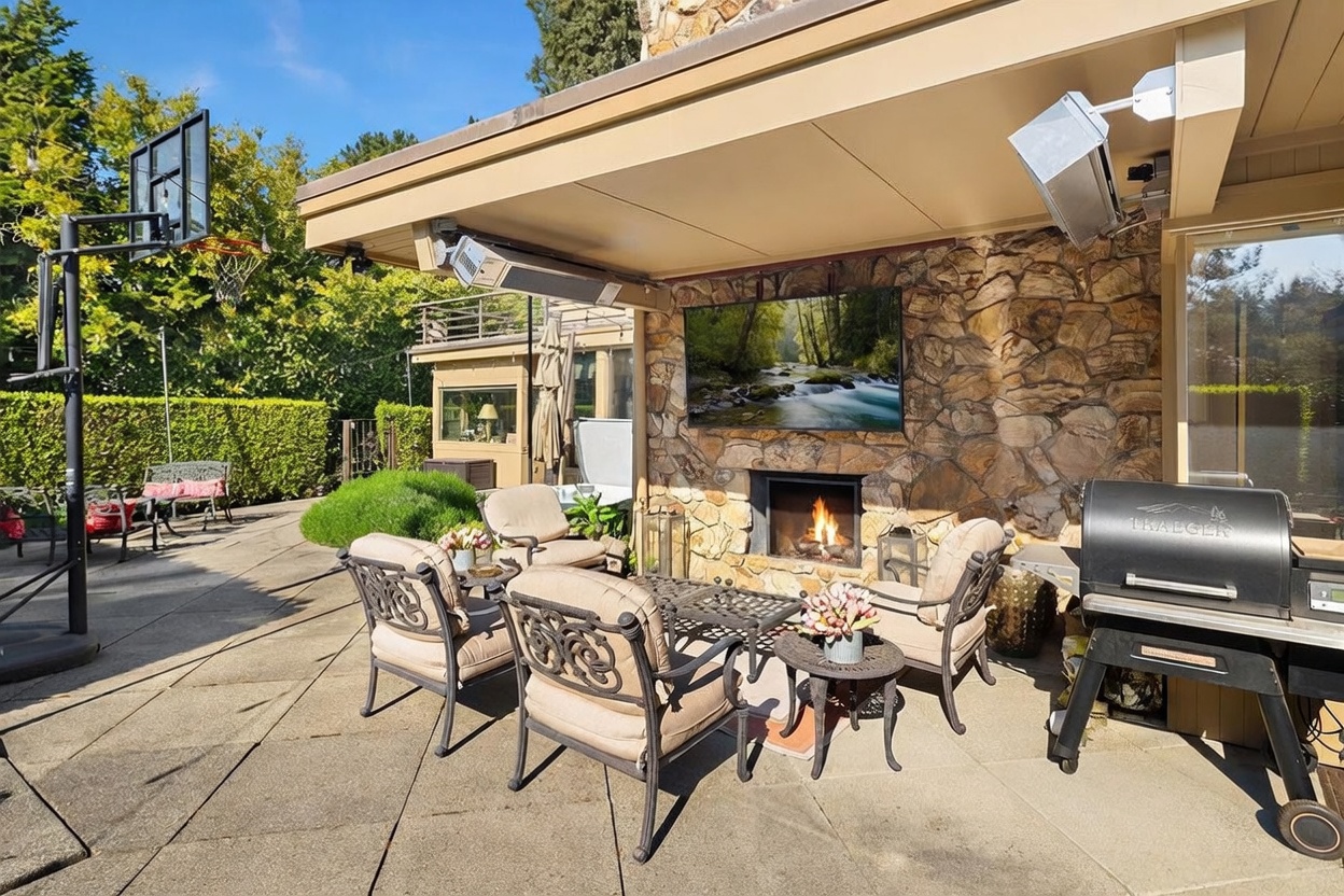 This inviting outdoor living space features a stone-clad chimney breast with an integrated fireplace and a mounted television, creating a cozy entertainment area. The patio is furnished with comfortable cushioned seating and a small side table, while a Traeger grill sits to the right. The scene is set under a covered patio roof, providing a perfect transition between the indoor living space and the surrounding backyard.
