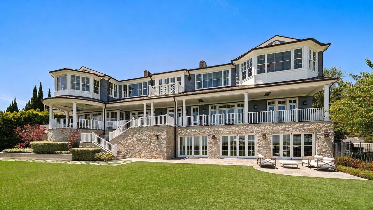 This expansive multi-story residence features a classic shingle-style exterior with a prominent stone-clad lower level. The rear facade showcases a large elevated deck with white railings, multiple sets of French doors leading to a stone patio, and a manicured lawn in the foreground. The architectural design emphasizes symmetry and traditional elegance, set against a clear blue sky.