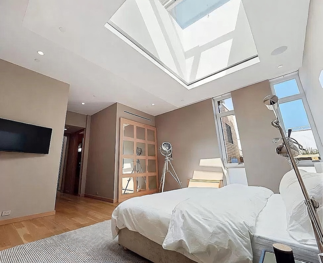 This modern primary bedroom features a large skylight that floods the space with natural light, highlighting the neutral-toned walls and light wood flooring. The room is furnished with a plush bed, a unique tripod-style floor lamp, and a large glass-paneled wardrobe. The perspective is from the foot of the bed, looking toward the corner of the room and the doorway, creating an airy and sophisticated atmosphere.
