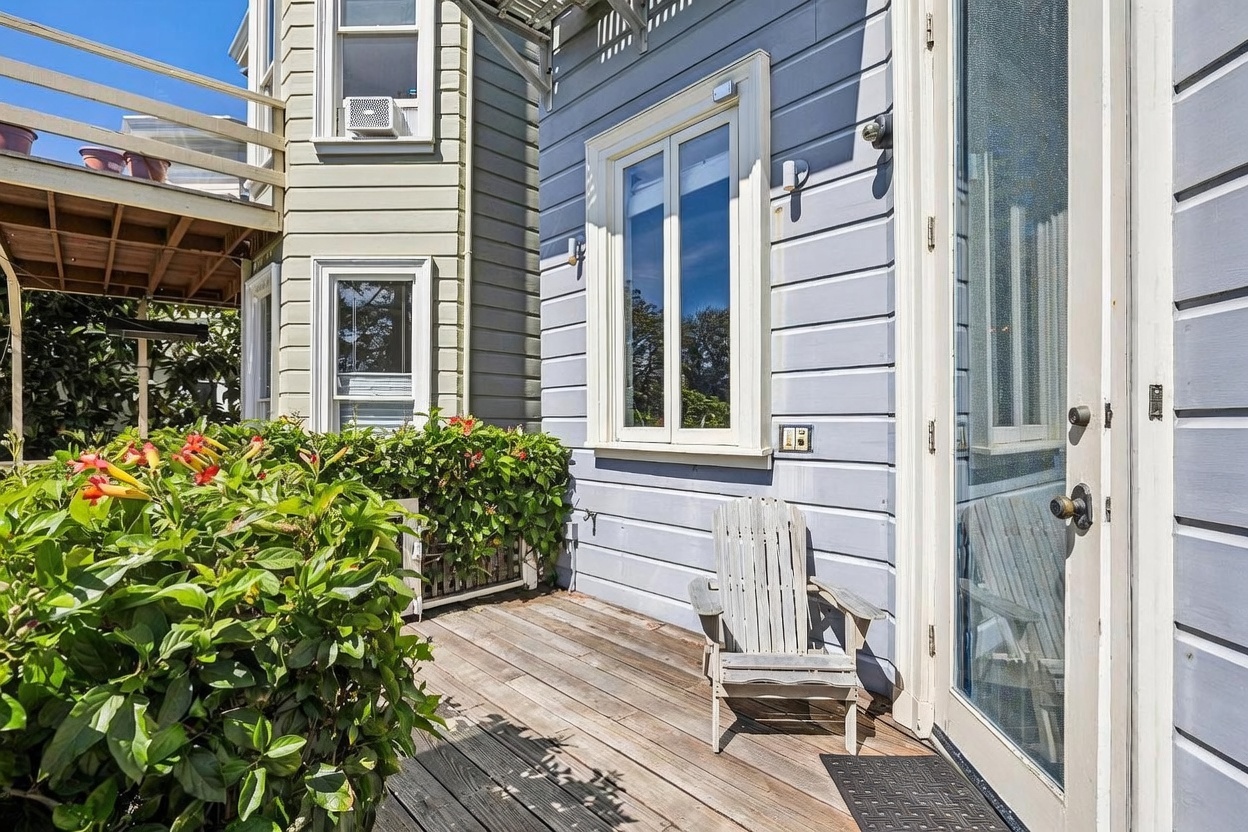 This image captures a cozy, sunlit wooden deck featuring a classic Adirondack chair positioned near a glass-paned door. To the left, a lush garden with vibrant red trumpet-shaped flowers adds a touch of natural color, while the light blue horizontal siding of the house provides a clean, coastal aesthetic. The perspective is a medium-wide shot from the deck surface, highlighting the inviting outdoor living space and the architectural transition between the deck and the home's exterior.