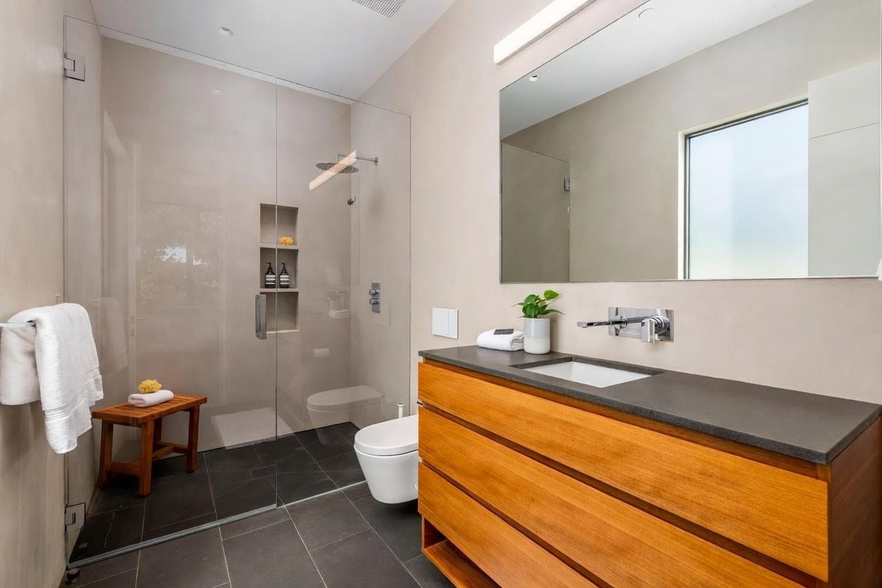 This modern bathroom features a sleek, wall-mounted vanity with a warm-toned wood finish and a dark stone countertop. A glass-enclosed walk-in shower with dark floor tiles and a built-in niche sits adjacent to a wall-mounted toilet. The space is illuminated by a large, frameless mirror and a minimalist light fixture, creating a clean and contemporary aesthetic.