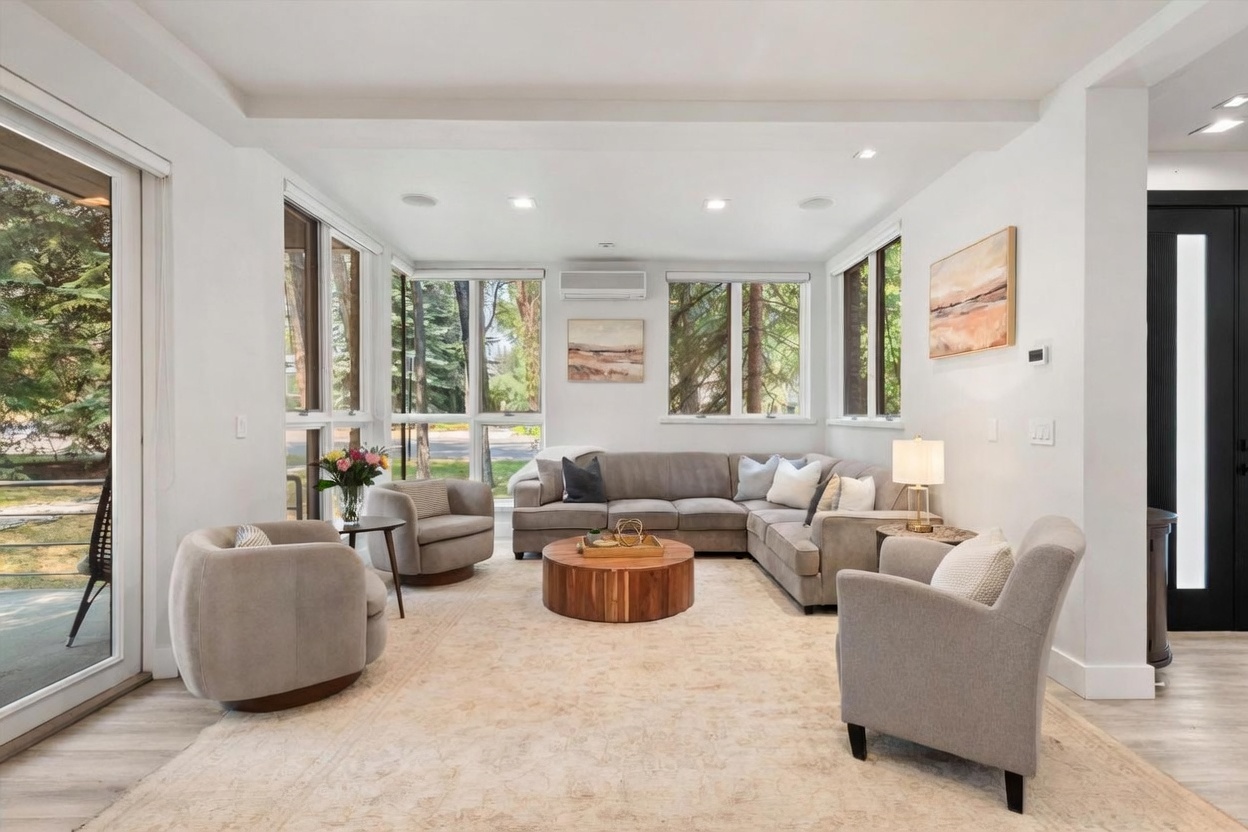 This bright and airy living room features a neutral color palette with a large sectional sofa, two swivel armchairs, and a central round wooden coffee table. The space is anchored by a large, light-toned area rug and benefits from abundant natural light provided by expansive windows and a wide glass door that opens to a lush, tree-filled outdoor area. The room has a clean, contemporary aesthetic with recessed lighting and a wall-mounted climate control unit.