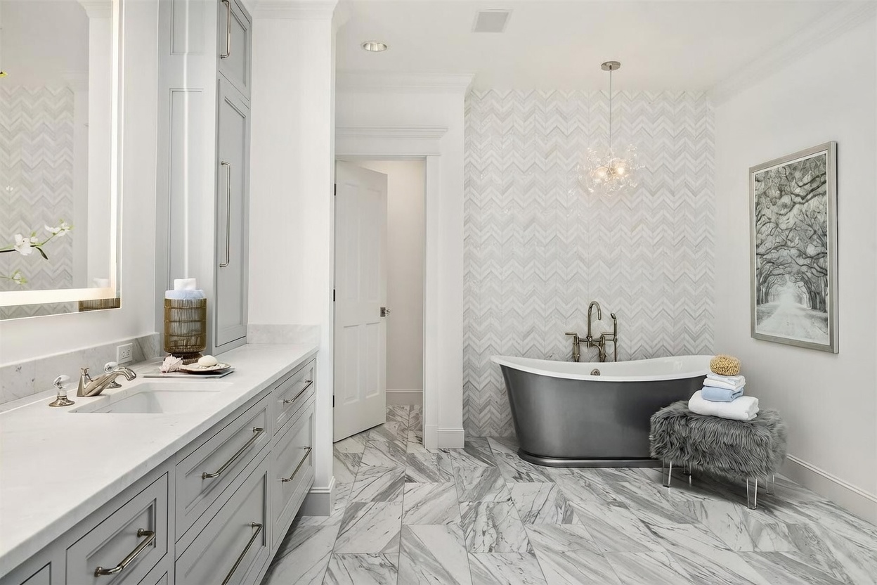 This luxurious primary bathroom features a striking freestanding dark-toned bathtub set against a wall with intricate herringbone-patterned tile. To the left, a long vanity with light-colored stone countertops and gray cabinetry provides ample storage, while a plush, furry stool sits beside the tub. The room is finished with large-format patterned stone flooring and a modern glass chandelier, creating a sophisticated and serene atmosphere.