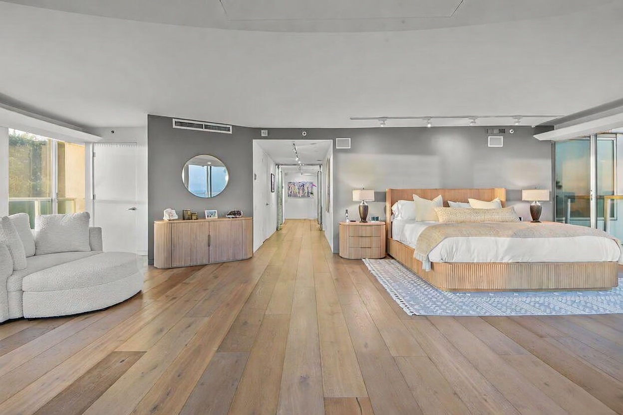 This spacious primary bedroom features light wood flooring and a neutral color palette, creating an airy and sophisticated atmosphere. The room is furnished with a large bed with a slatted headboard, a matching sideboard, and a curved lounge chair, all arranged to emphasize the open floor plan. A long hallway leads away from the sleeping area, while large windows provide ample natural light and a view of the outdoors.