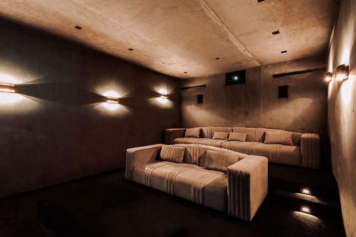 This sophisticated home theater features a tiered seating arrangement with two plush, modern sofas upholstered in a neutral fabric. The room is characterized by its moody, monochromatic aesthetic, enhanced by warm, directional wall sconces that create a cinematic atmosphere. The minimalist design and recessed lighting contribute to an intimate, high-end viewing experience.