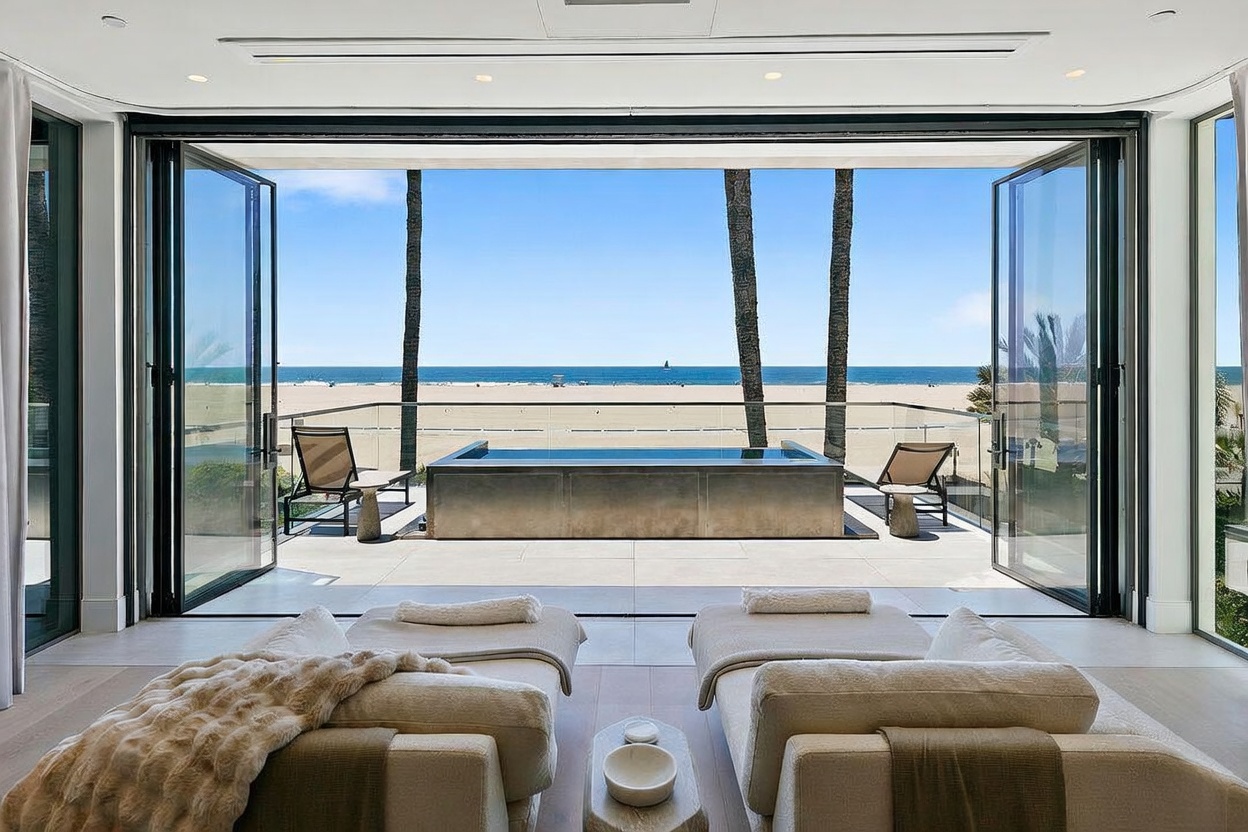 This luxurious interior space features two plush lounge chairs facing a wide-open, floor-to-ceiling glass wall that seamlessly connects the room to an outdoor terrace. The terrace showcases a sleek, rectangular plunge pool overlooking a pristine sandy beach and the ocean horizon. The overall aesthetic is one of sophisticated coastal elegance, emphasizing a fluid transition between indoor comfort and the expansive natural scenery.