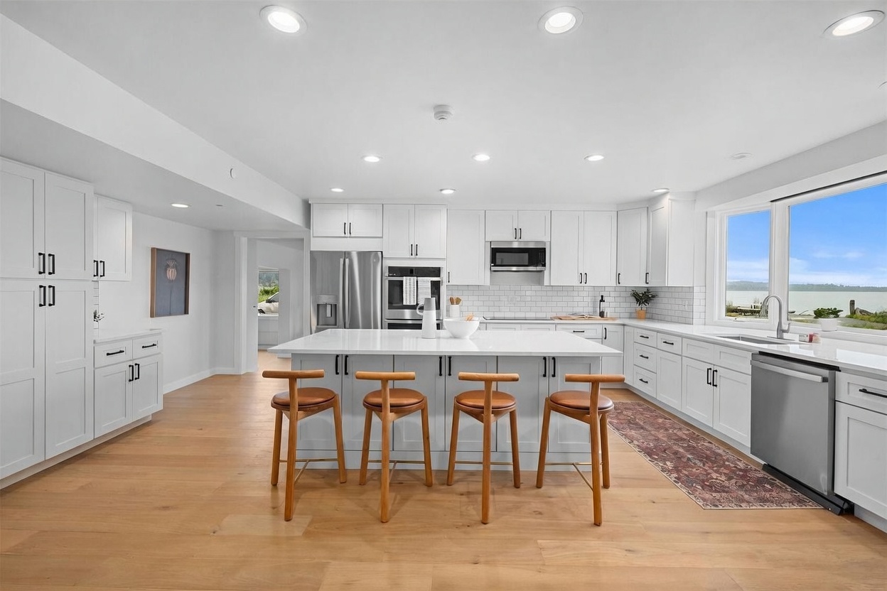 This bright, modern kitchen features white cabinetry, a large central island with seating for four, and light-toned wood flooring. A large window above the sink provides a scenic view of the water, while recessed lighting and pendant lights illuminate the workspace. The space is clean and airy, showcasing a functional layout with stainless steel appliances and a patterned runner rug.