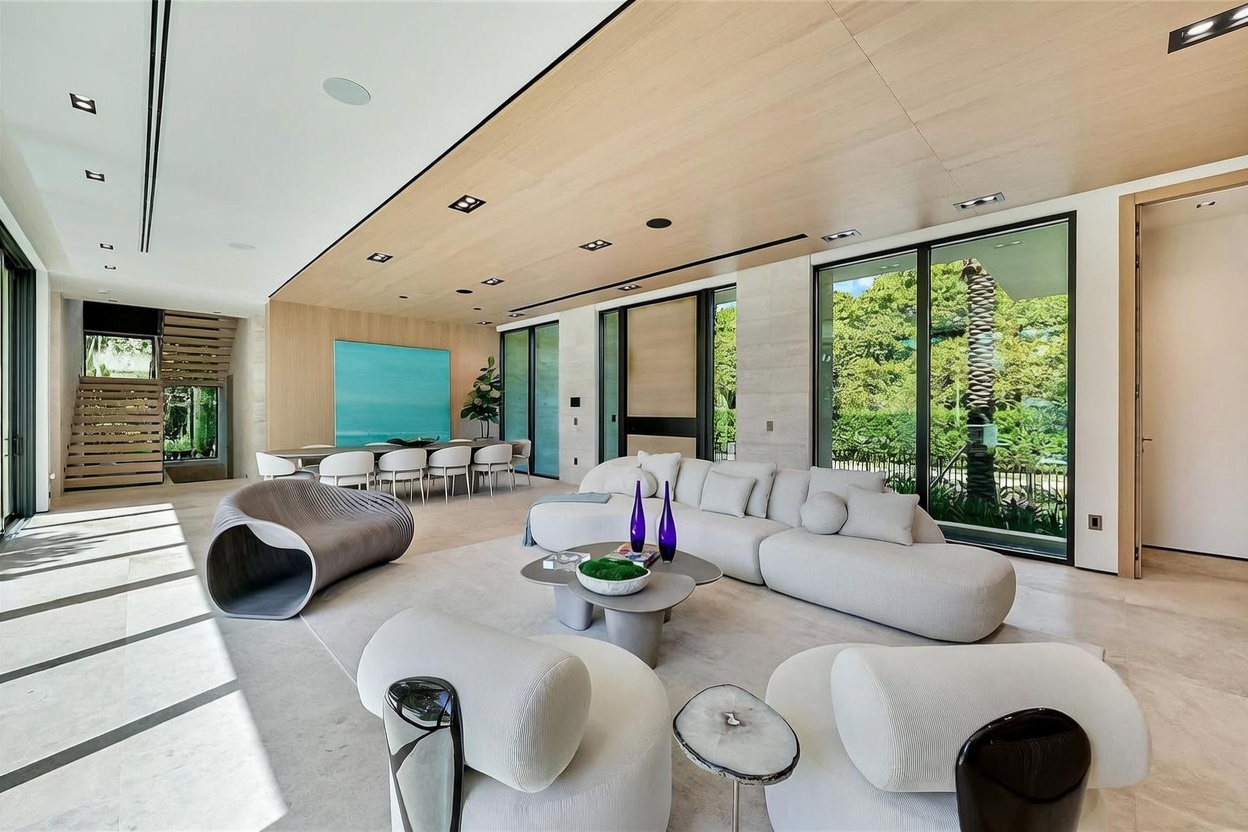 This expansive, open-concept living area features a contemporary design with light-toned stone flooring and a warm wood-paneled ceiling. The space is furnished with a large, curved white sectional sofa, a unique sculptural lounge chair, and a central coffee table, all oriented toward the bright, airy atmosphere created by large windows overlooking lush greenery. A dining area is visible in the background, anchored by a vibrant blue abstract painting, while a staircase with floating treads adds a modern architectural element to the side.