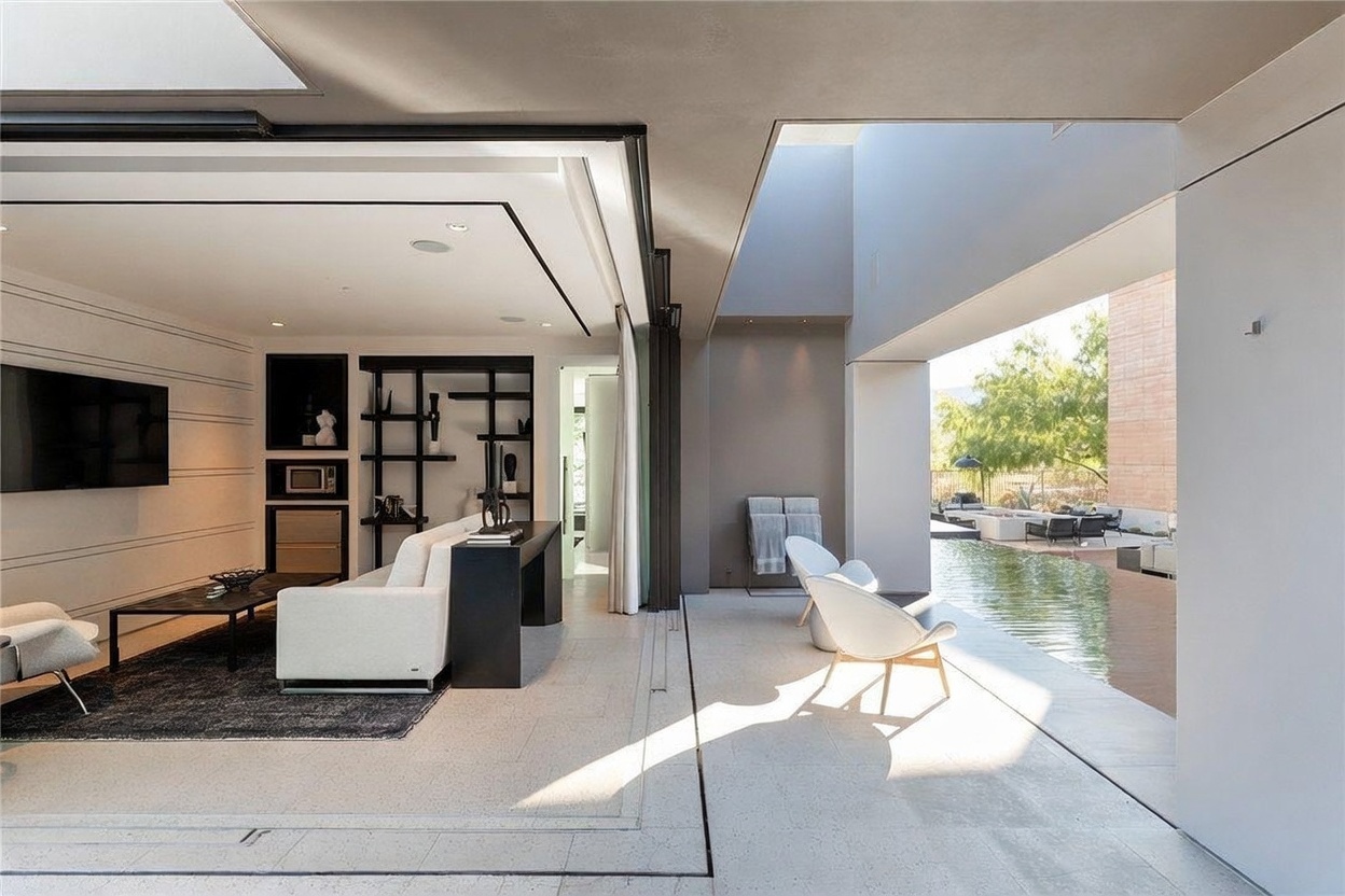 This sophisticated indoor-outdoor living space features a seamless transition between a modern living room and a covered patio area overlooking a pool. The interior includes a white sofa, a dark area rug, and a minimalist black shelving unit, while the exterior space is furnished with sleek white lounge chairs. The open-concept design is defined by large glass panels that slide away to merge the two areas, creating an airy and luxurious atmosphere.