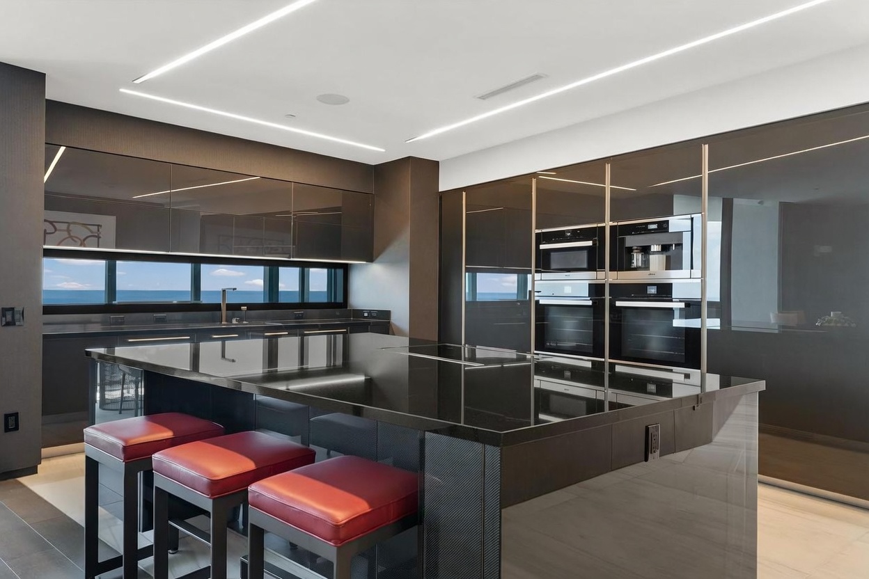 This sleek, modern kitchen features high-gloss dark cabinetry and a large central island with a dark stone countertop, complemented by three vibrant red bar stools. The space is illuminated by integrated linear lighting and includes built-in stainless steel appliances, such as dual ovens and a coffee system. A horizontal window strip provides a panoramic view of the ocean, enhancing the sophisticated and minimalist aesthetic of the room.