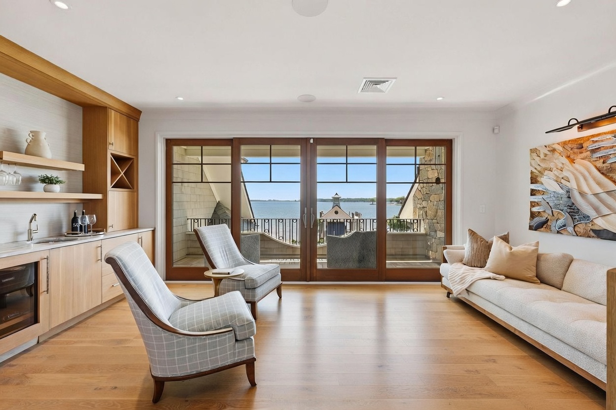This bright and airy living space features a wet bar with light wood cabinetry and open shelving, seamlessly transitioning into a comfortable seating area. Large glass doors provide an expansive view of the water and a private balcony, filling the room with natural light. The decor includes a neutral-toned sofa, patterned armchairs, and a textured rug, creating a sophisticated and inviting atmosphere.