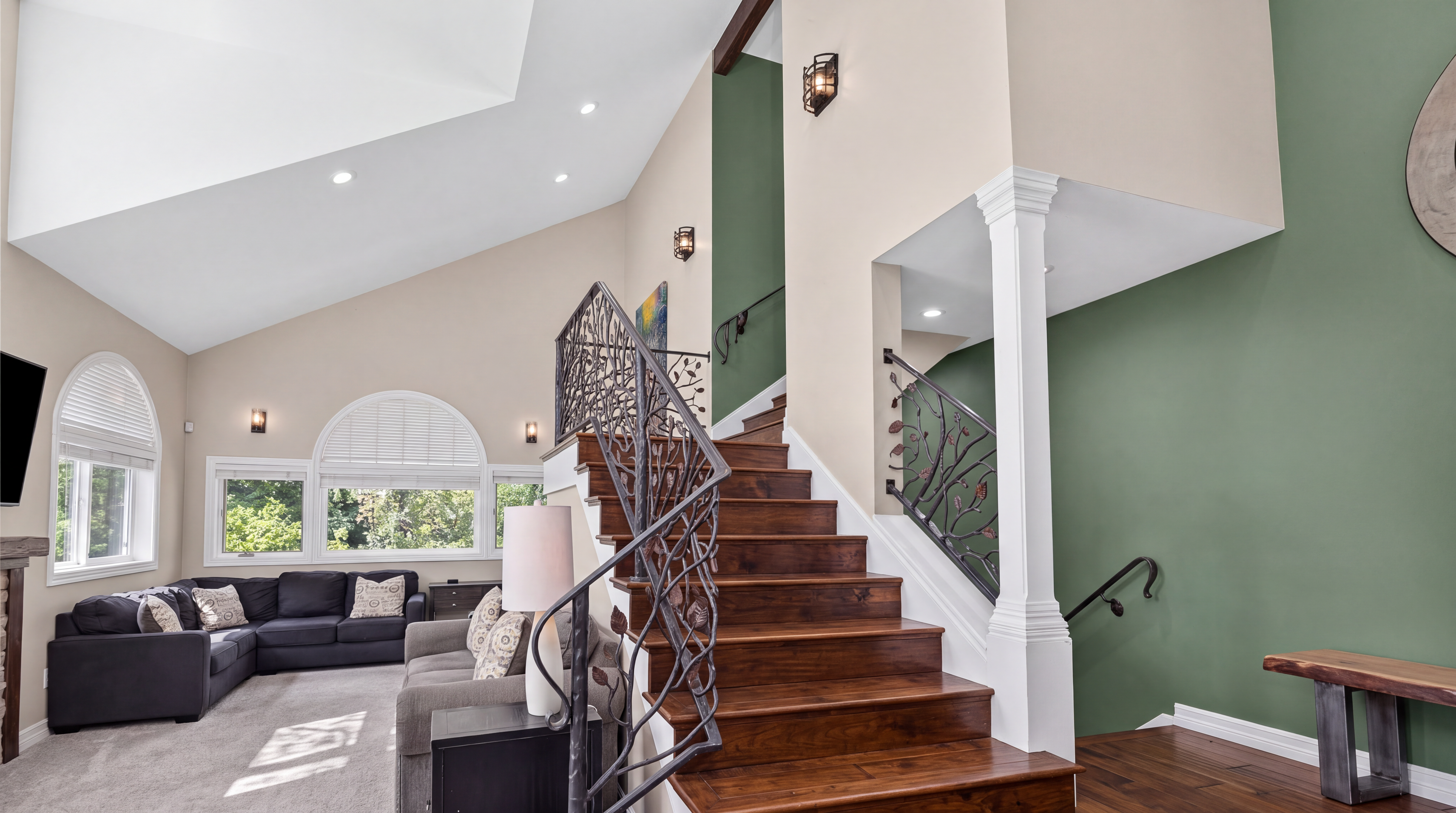 This grand foyer features a striking wooden staircase with intricate wrought-iron railings that serve as a focal point. To the left, an open-concept living area provides a cozy space with vaulted ceilings and abundant natural light from arched windows. The home utilizes a bold color palette, contrasting neutral walls with deep green accents and rich hardwood flooring for a sophisticated, high-end feel.