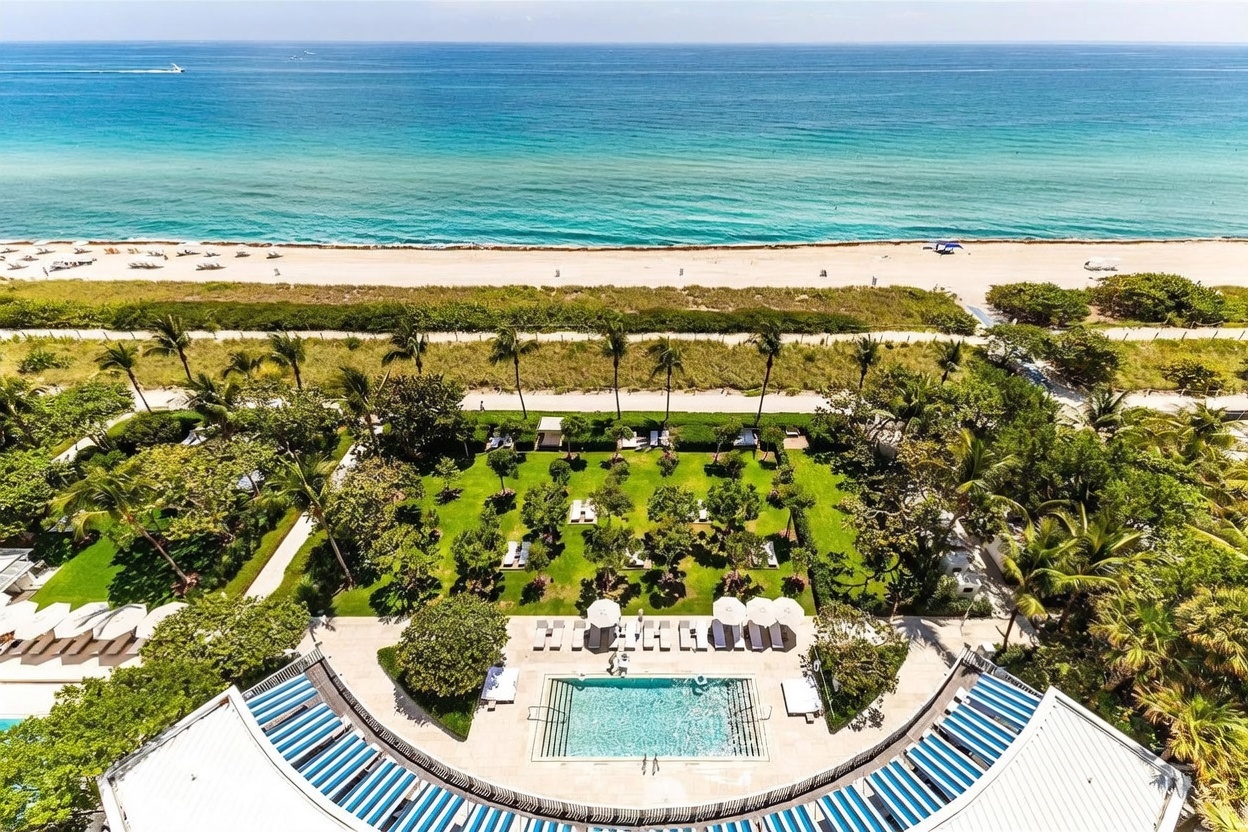 This high-angle aerial view captures a luxurious beachfront property featuring a curved swimming pool surrounded by a manicured lawn and lush tropical landscaping. Beyond the private garden, a paved walkway separates the property from the sandy beach and the expansive blue ocean. The perspective emphasizes the seamless transition from the private residence to the coastal environment, highlighting the prime waterfront location.